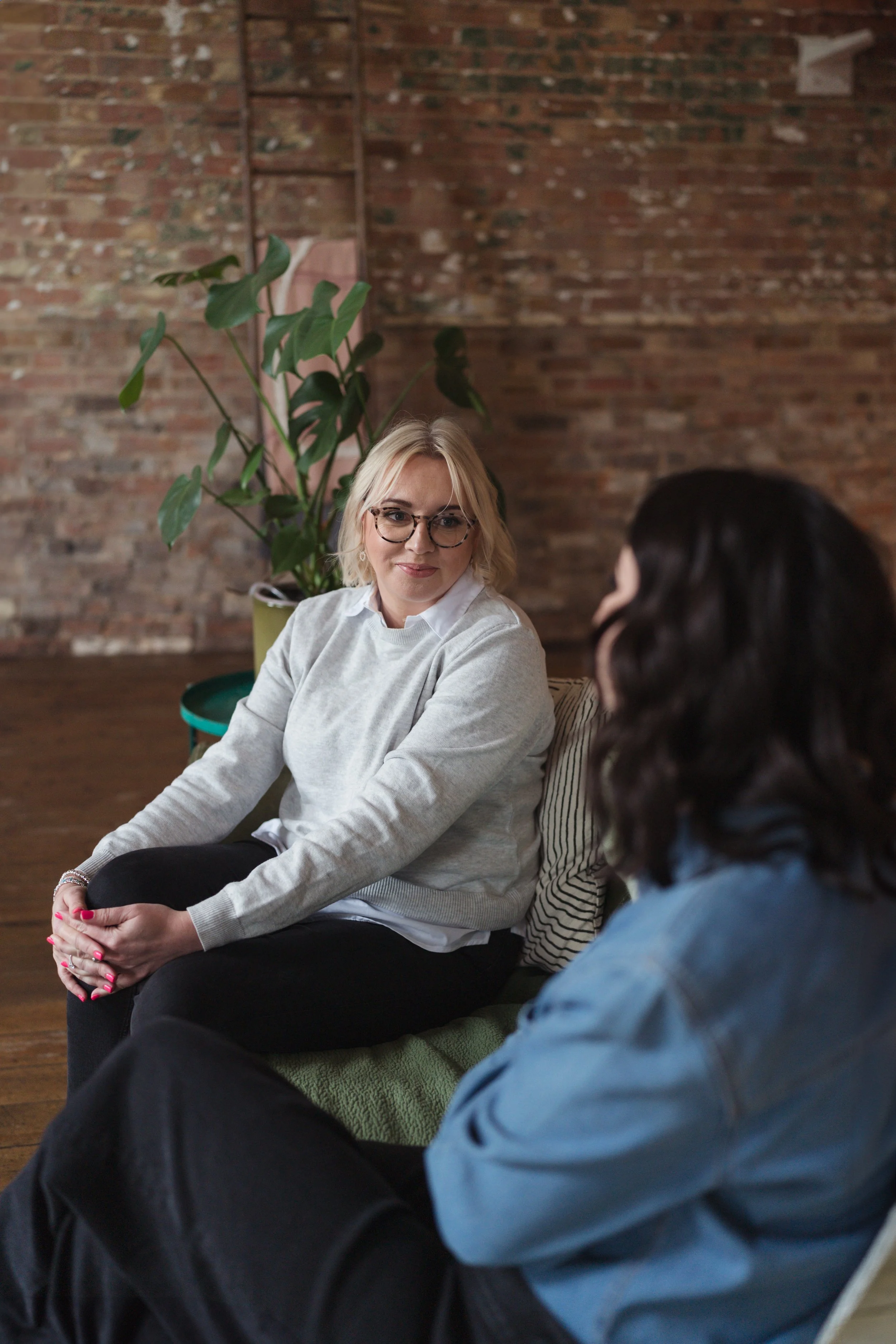 Two women are having a conversation in a cozy indoor setting.  Sadie Finch, founder of More Than Admin, smiles while sitting on a couch, facing a client with her back to the camera. A large green leafy plant and a brick wall are in the background.