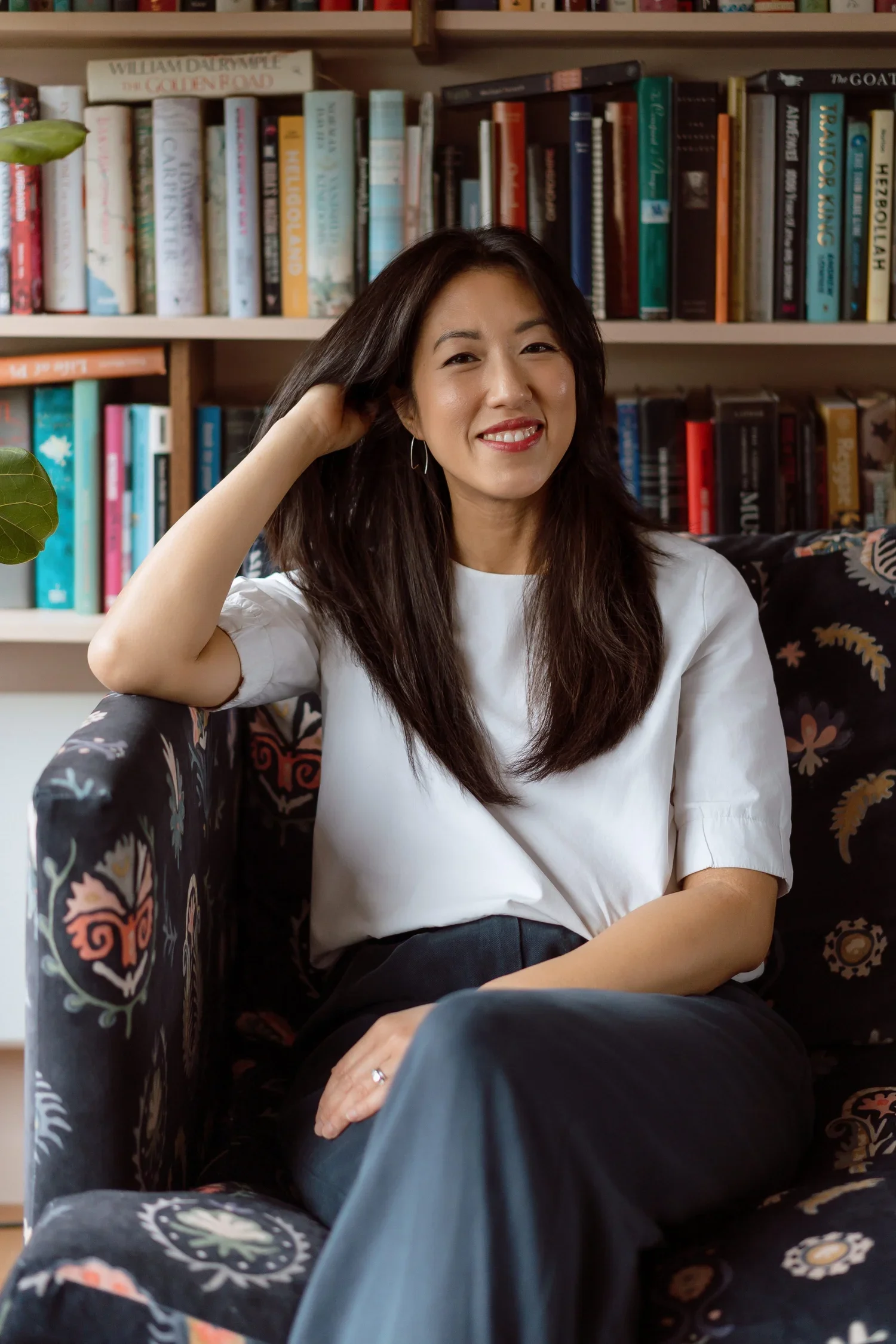 A woman with long dark hair, wearing a white top, sitting on a patterned armchair in front of a bookshelf filled with books, smiling at the camera.