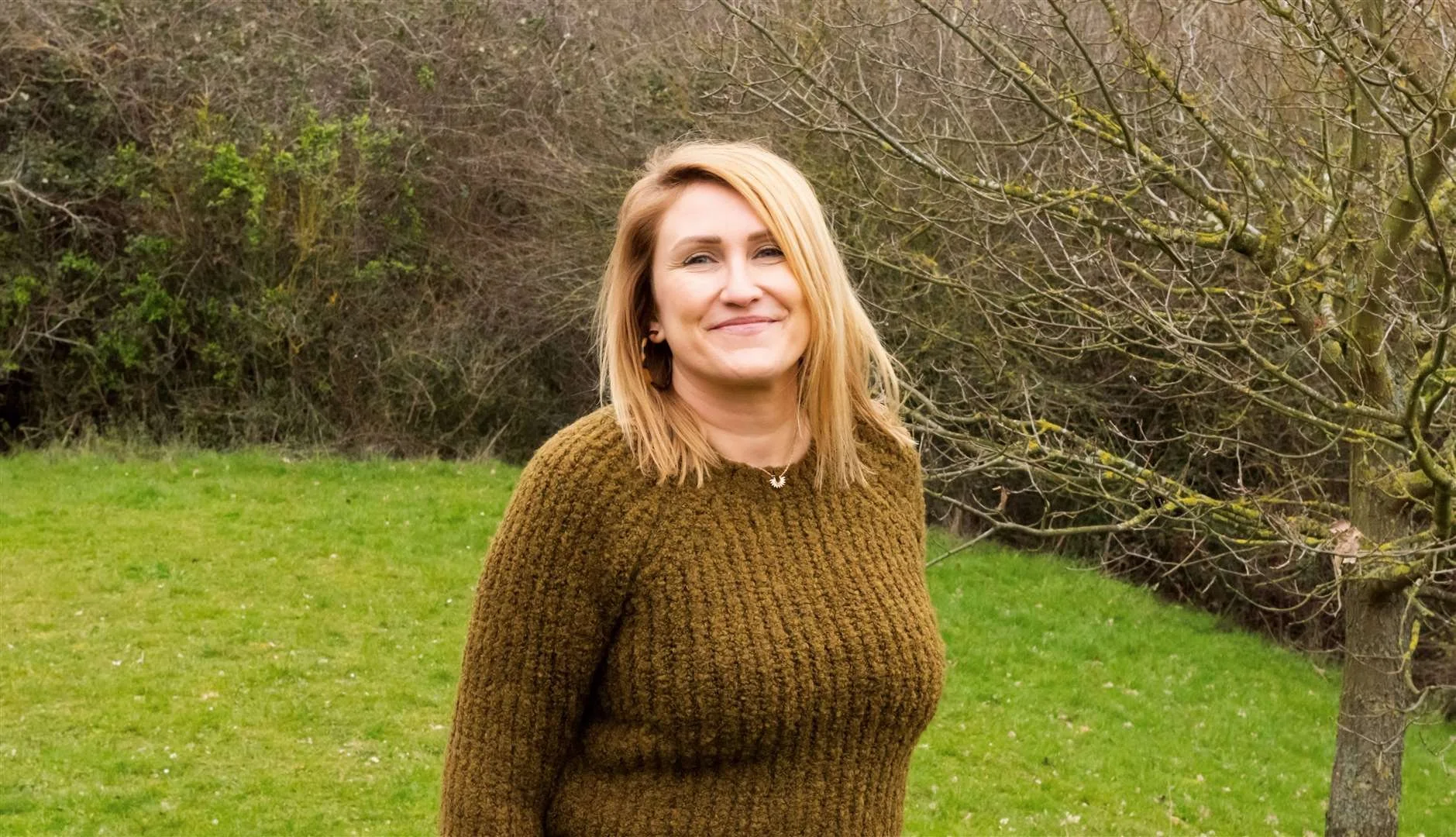 A woman with shoulder-length blonde hair, wearing a brown knitted sweater, standing outdoors on a grassy field with trees and bushes in the background.