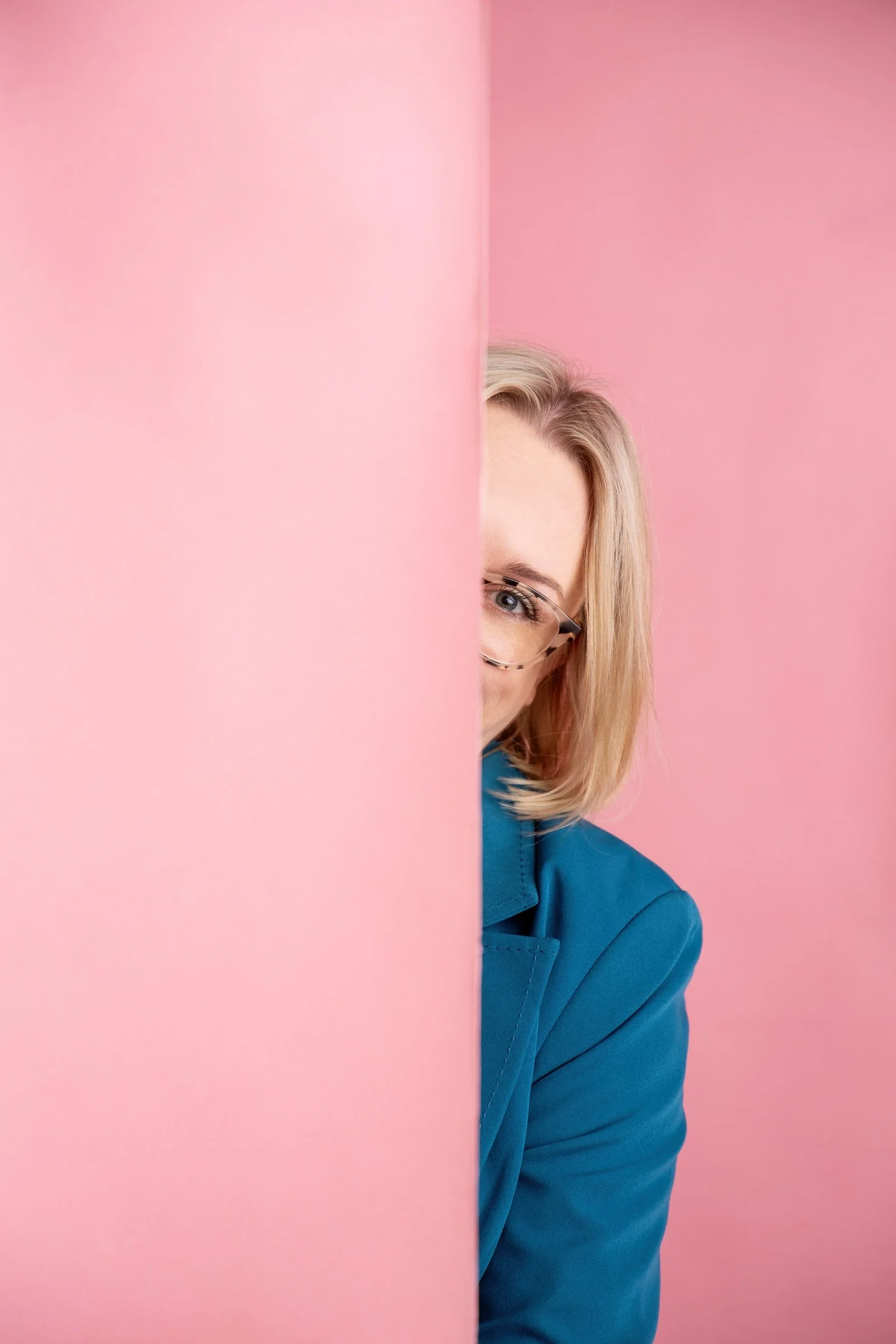 Sadie Finch, founder of More Than Admin ( woman with blonde hair, glasses) and a blue blazer peeking from behind a pink wall, smiling.