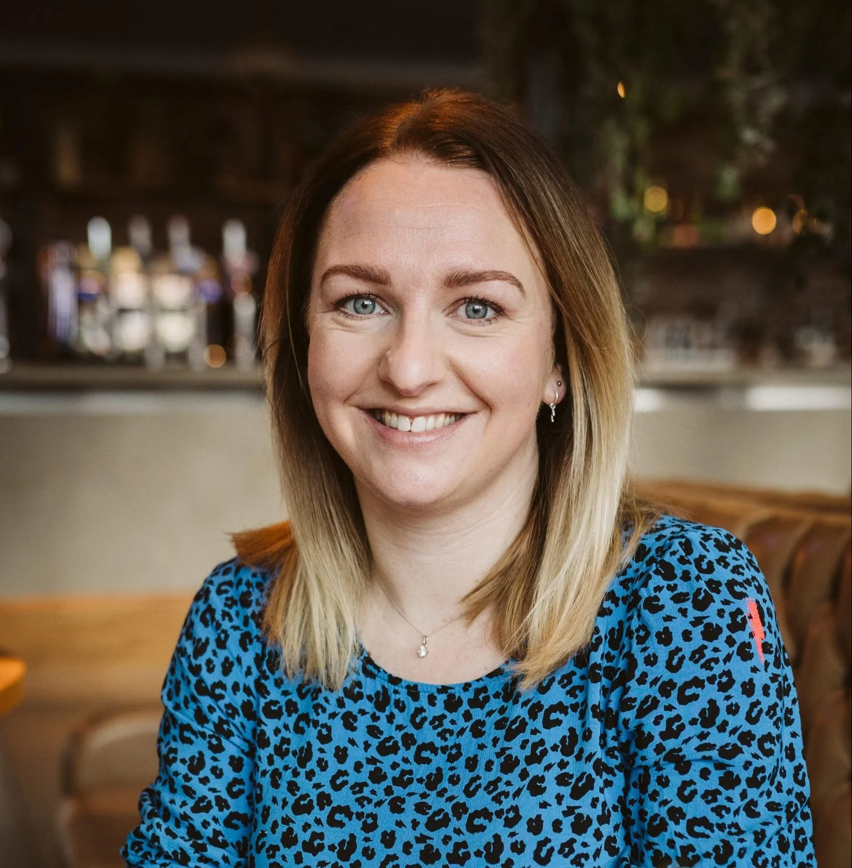 A woman with blonde hair and blue eyes, smiling in a casual setting with a blurred background of a bar or restaurant interior.