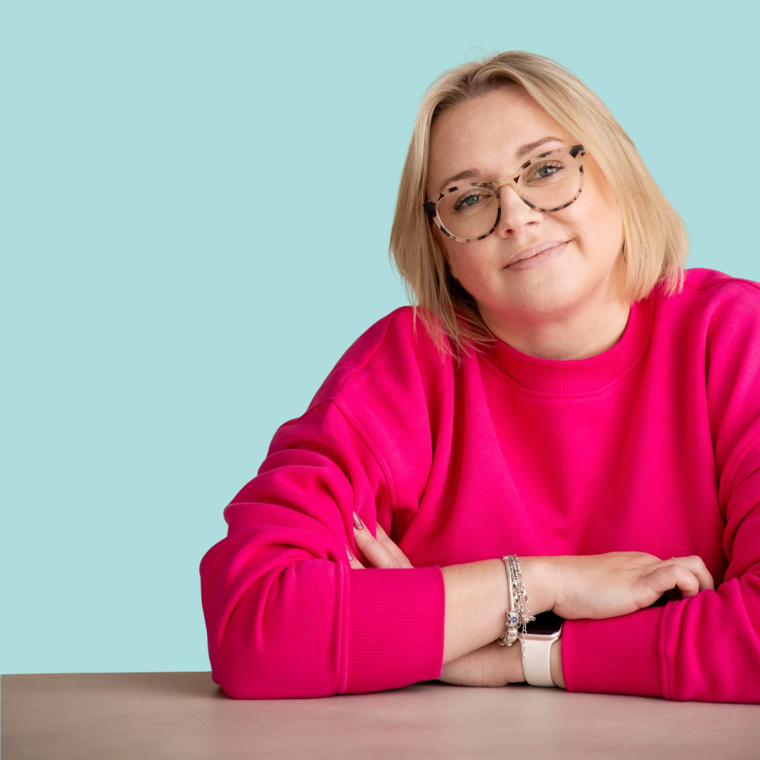 Sadie Finch with blonde hair, glasses, wearing a bright pink sweatshirt, seated at a table with her arms crossed, smiling slightly against a light blue background.