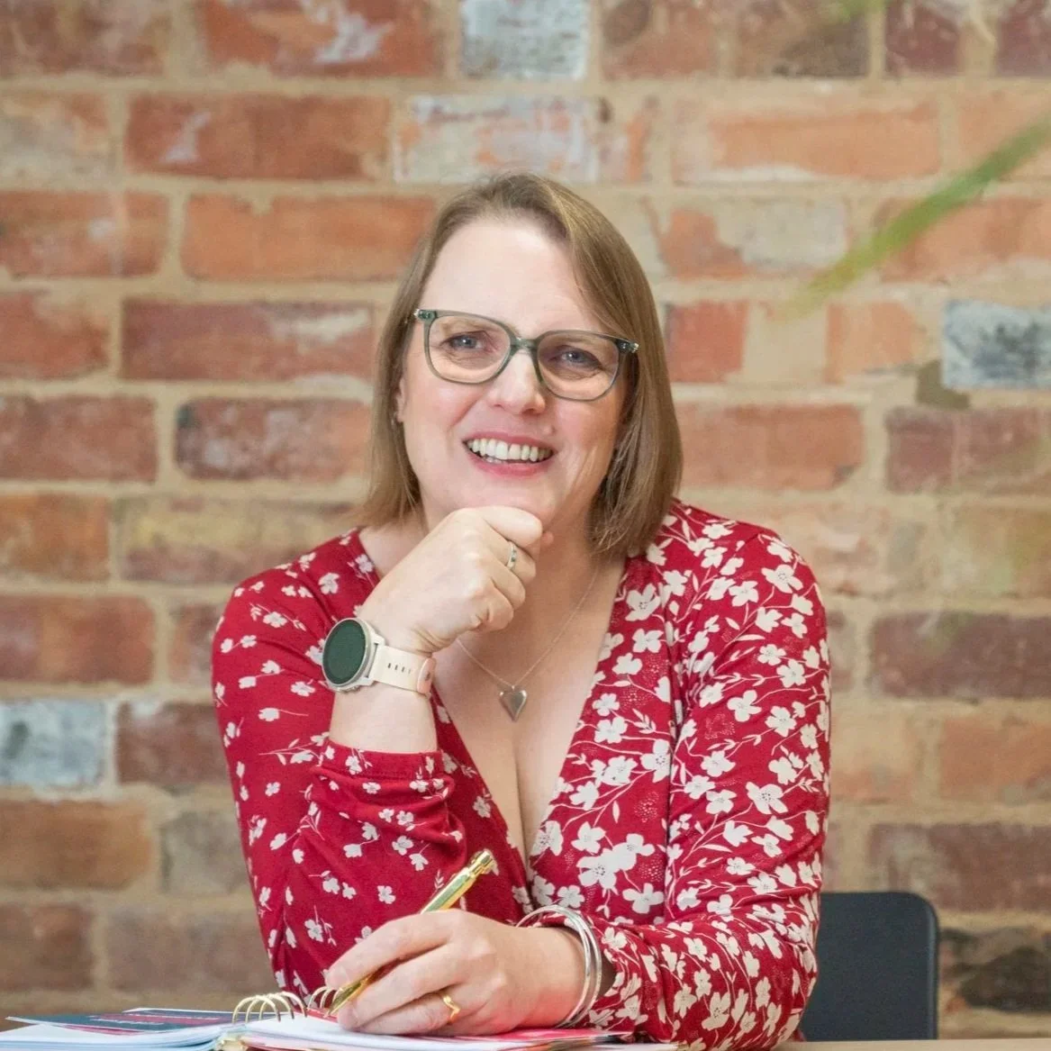 A smiling woman with glasses and shoulder-length brown hair, wearing a red floral blouse, sitting at a table with a spiral notebook and a pen, in front of a brick wall.