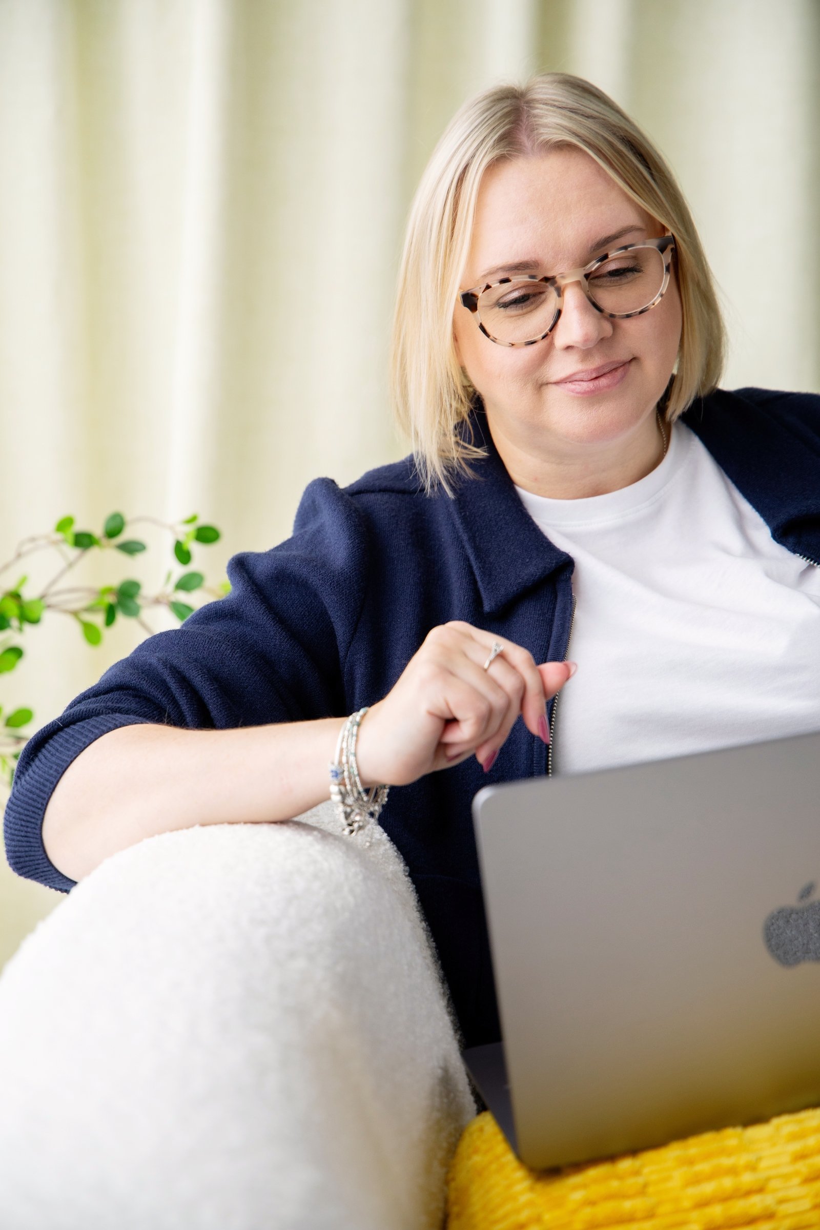 Sadie Finch, founder of More Than Admin  using a gray laptop, sitting on a cream-colored sofa with a yellow cushion, in a room with light-colored curtains and a plant in the background.
