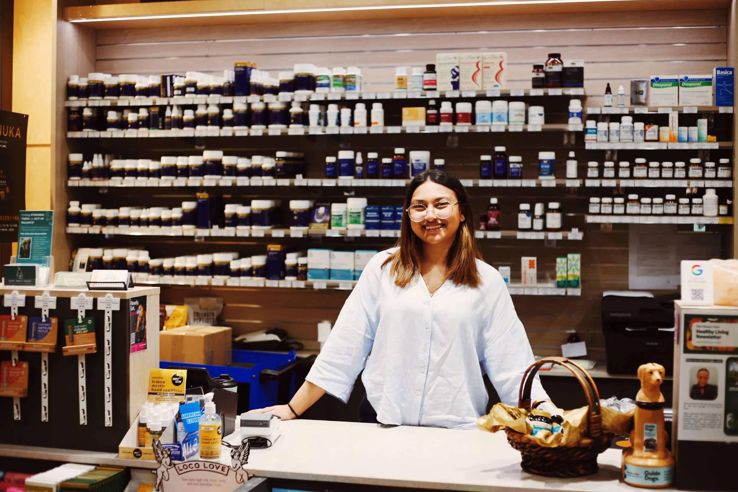 A woman with glasses smiling behind a checkout counter in a pharmacy or health store, with shelves of bottles and boxes of medication and health products in the background.