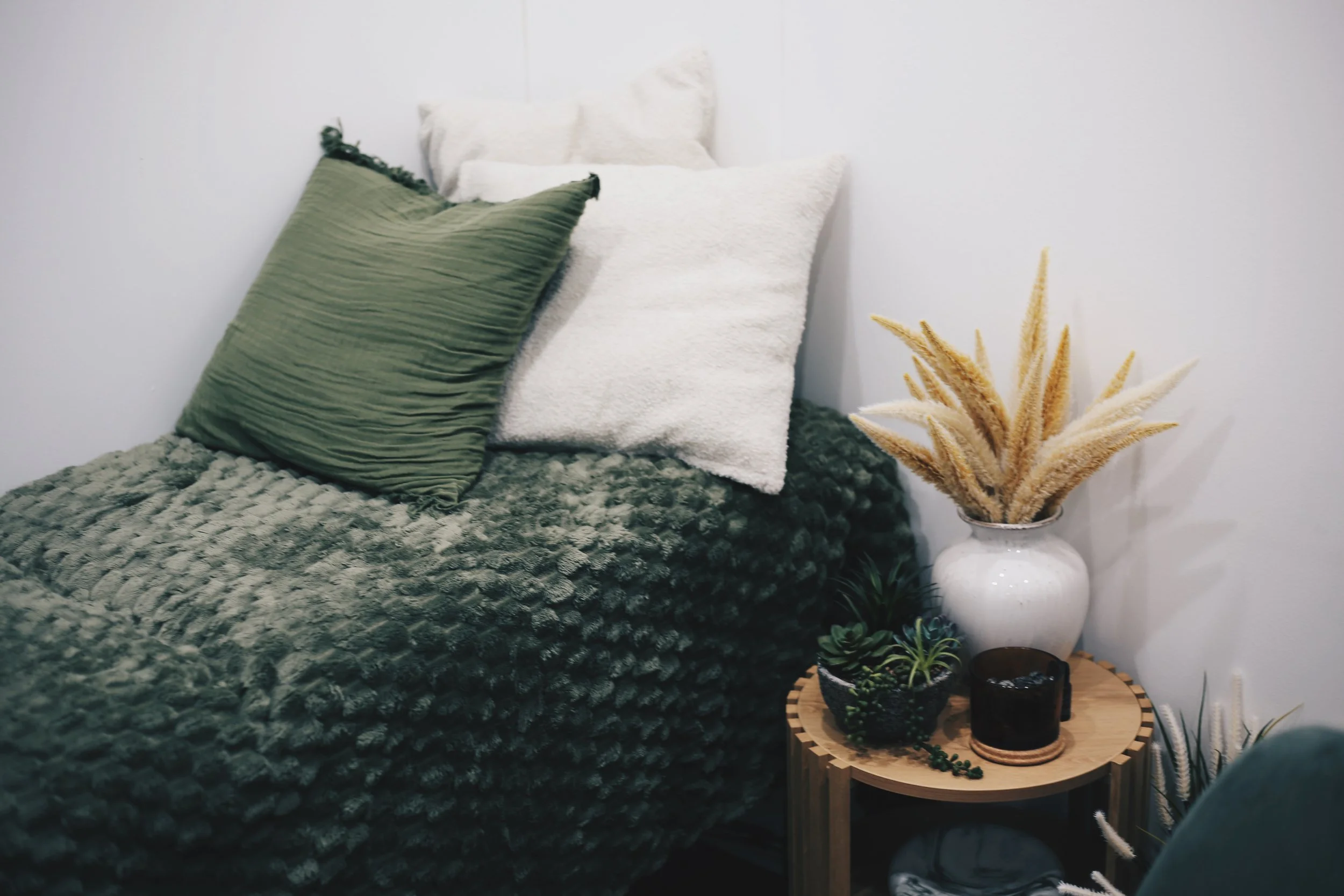 A cozy bedroom corner with a textured gray bedspread, a green plisse pillow, and white pillows, next to a side table with dried pampas grass in a white vase and potted plants on a round wooden tray.