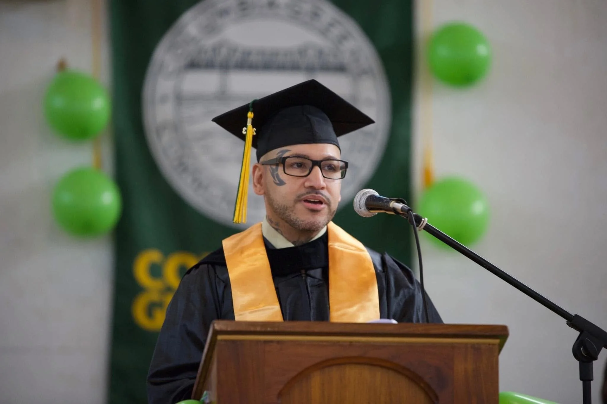 A man in graduation attire, wearing an academic cap and gown, standing behind a podium and speaking into a microphone.