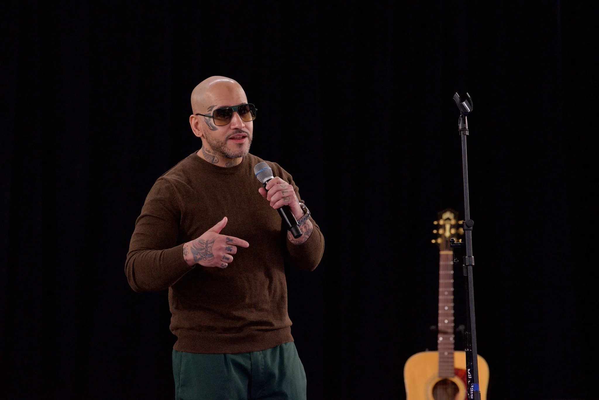 A bald man with tattoos and sunglasses holding a microphone, speaking on a stage with a guitar and microphone stand behind him.