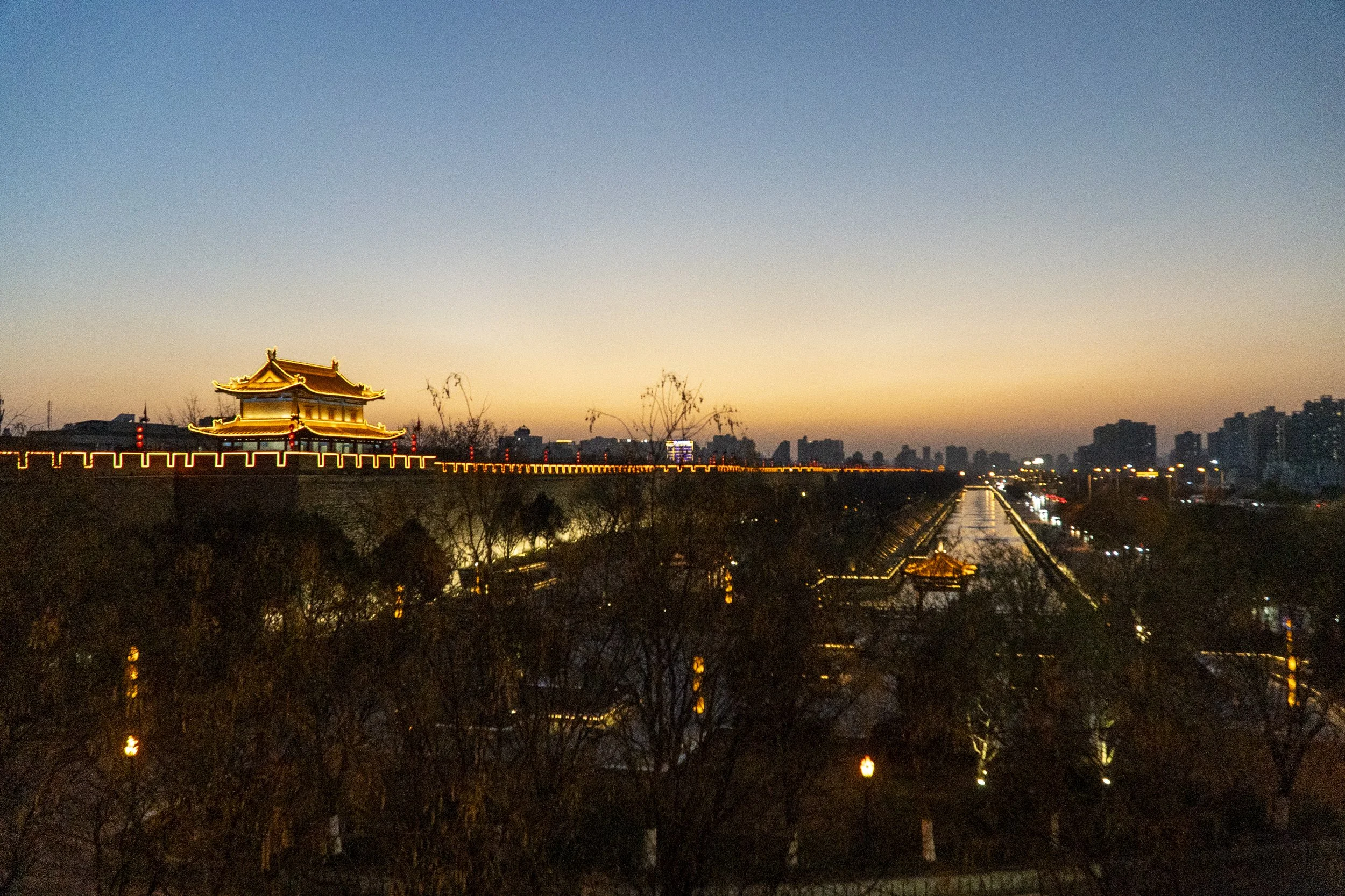 Sunset over a city with a traditional Chinese building illuminated on the left, a river with lights reflecting, and distant high-rise buildings on the right.