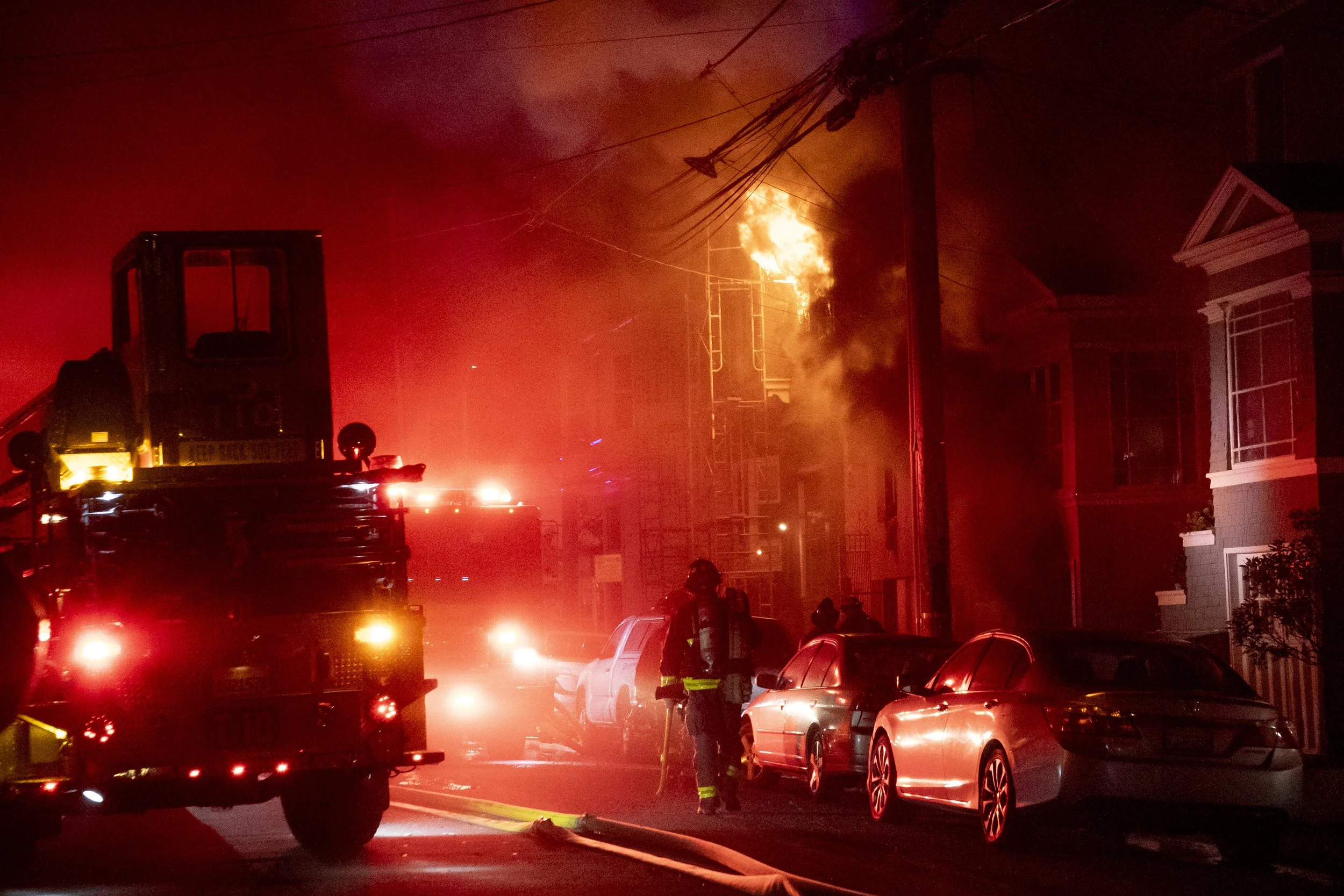 Firefighters respond to a building fire at night, with flames and smoke visible coming from the upper part of the building. Fire trucks and parked cars line the street, illuminated by red emergency lights.