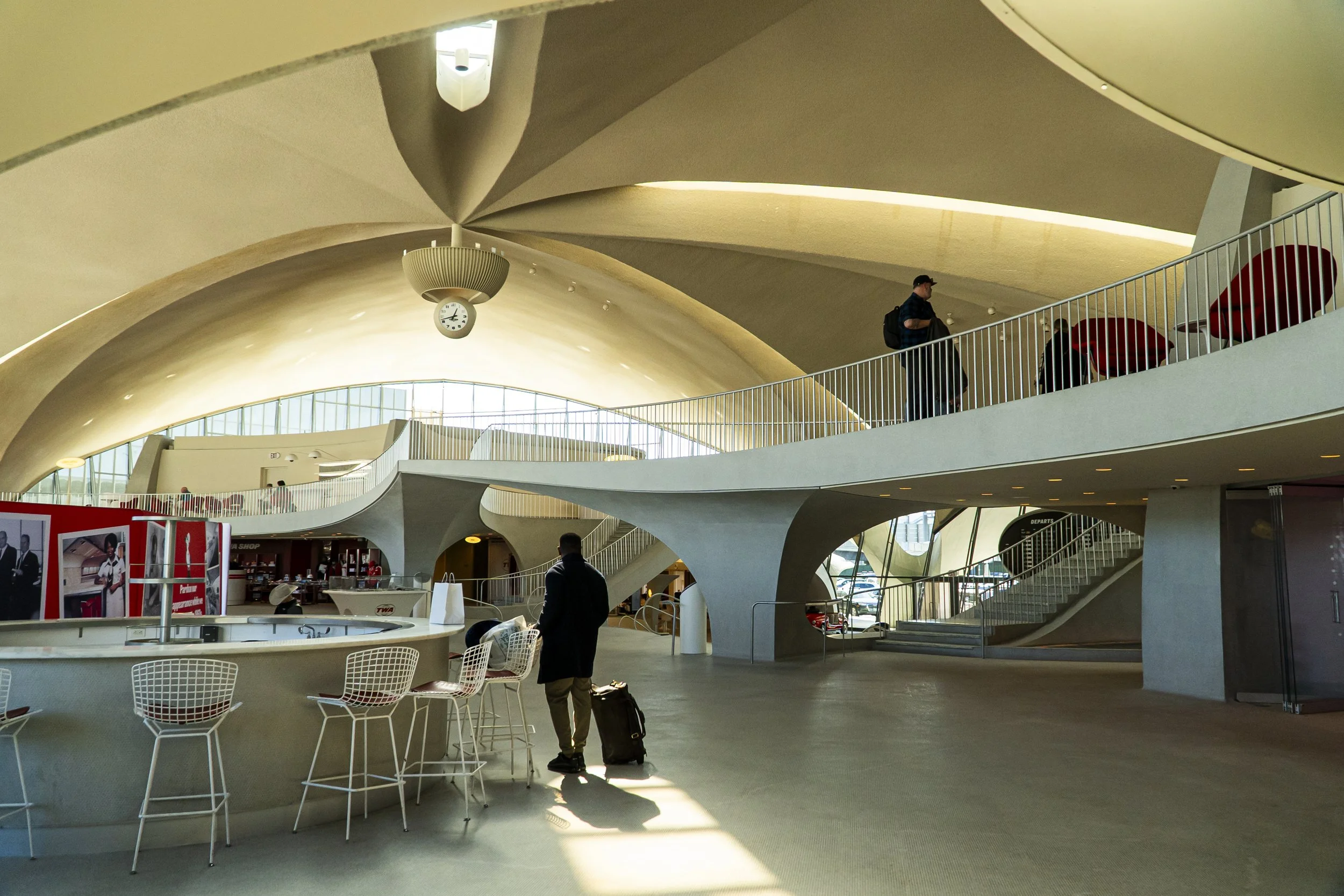Interior of an airport with modern architecture, featuring curved ceilings, large windows, a clock, and travelers with luggage.