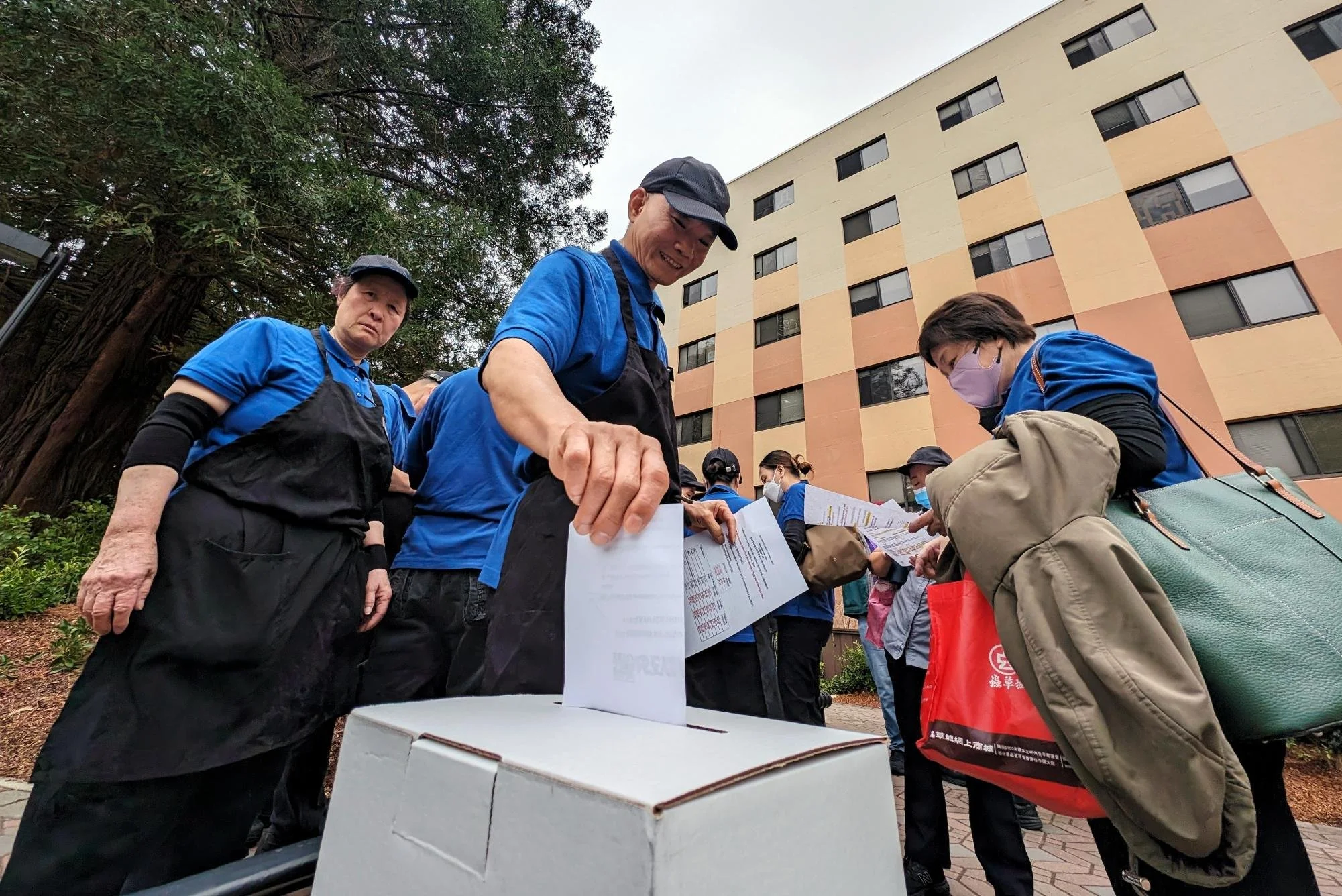 People in blue shirts and aprons casting ballots into a ballot box outside a building.