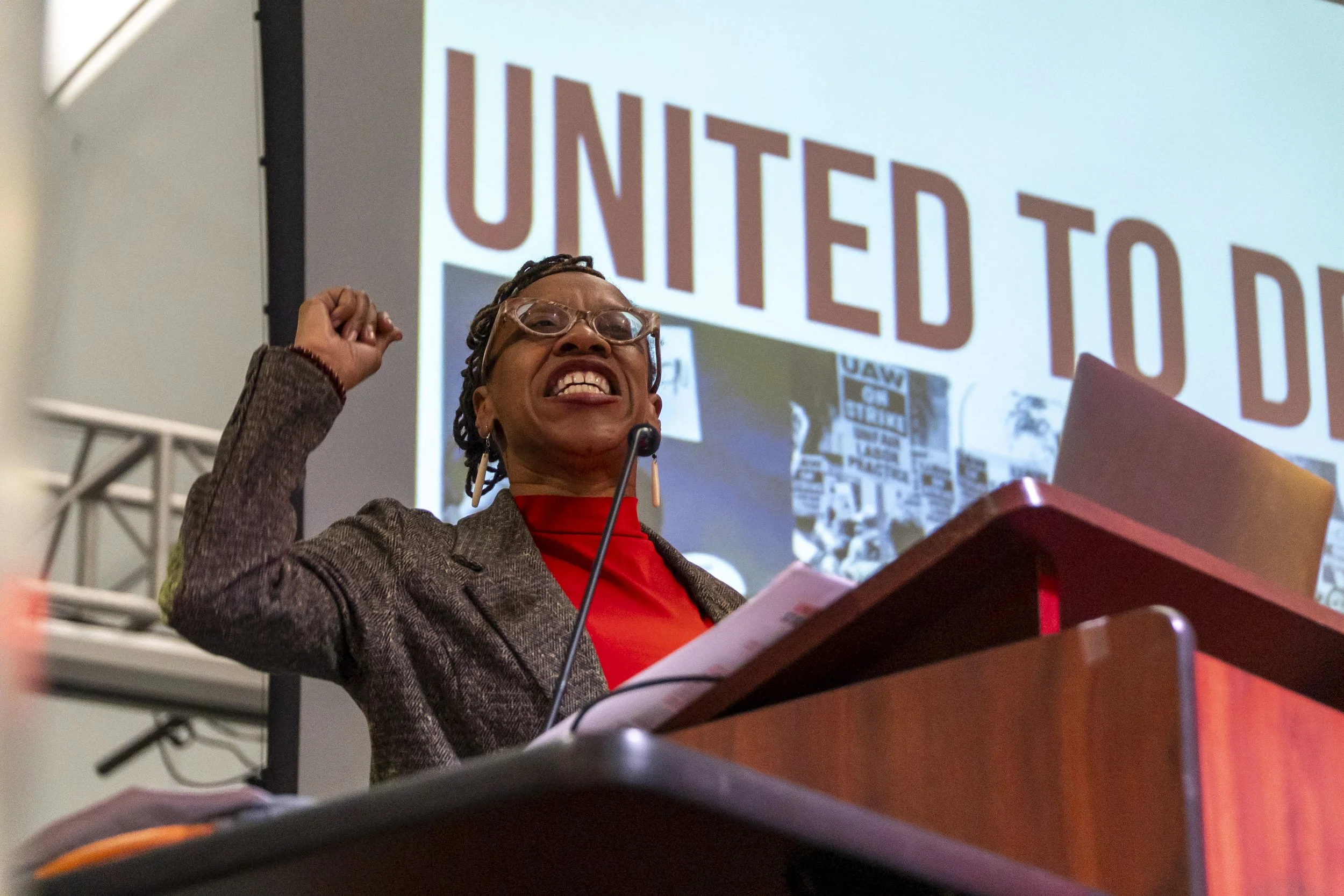 A woman wearing glasses, a red shirt, and a blazer stands at a podium, raising her right fist and smiling confidently in front of a large screen that says 'United to D' with people holding signs in the background.