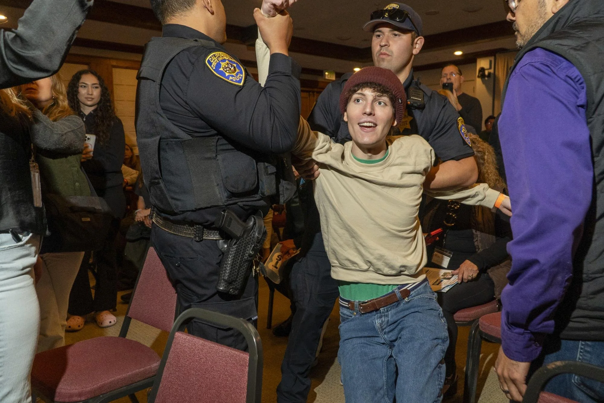 A young man in casual clothing and a beanie being detained or restrained by police officers, with people observing and some recording in the background.