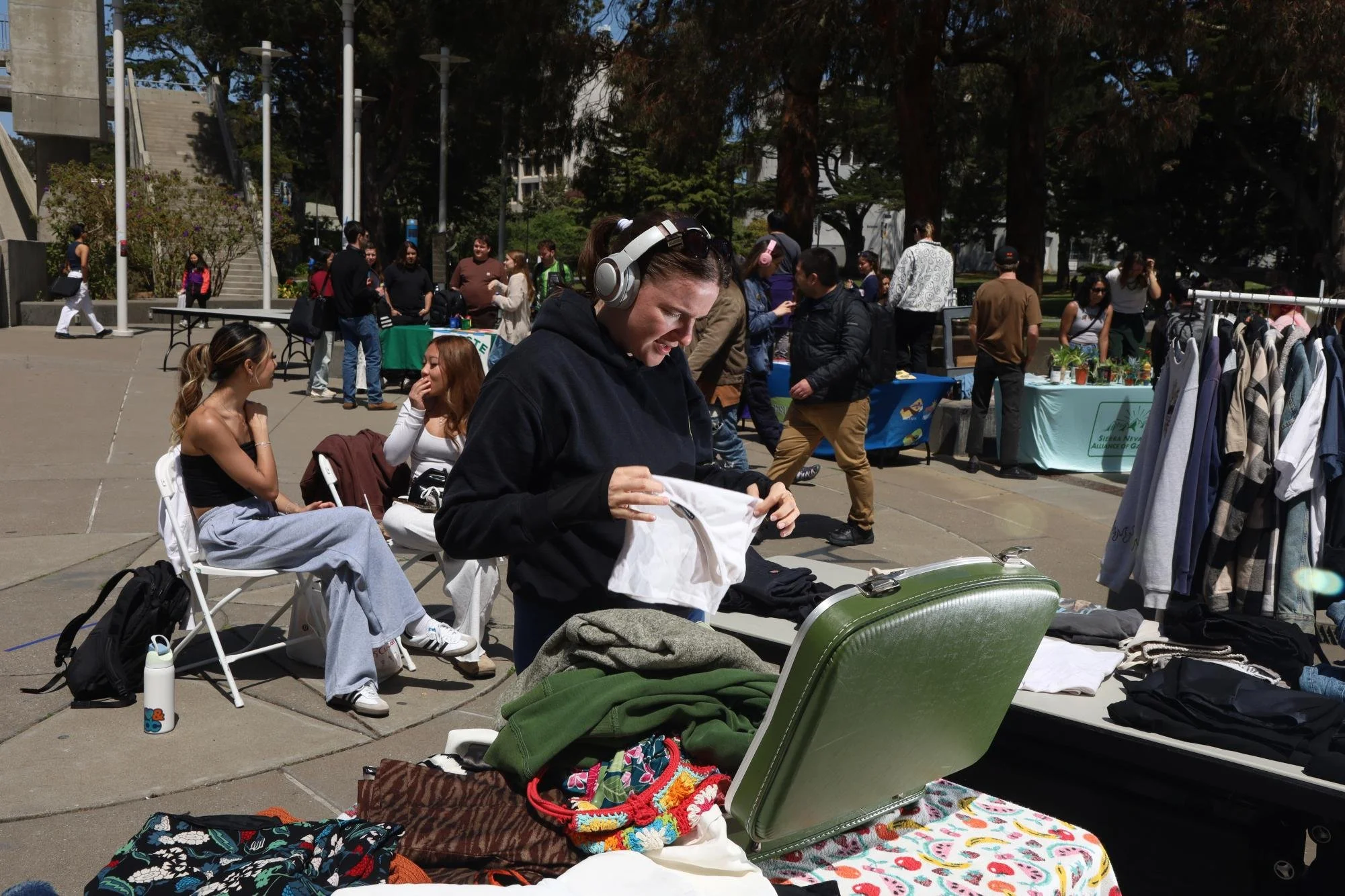 A woman wearing headphones is looking through a clothing item at an outdoor flea market. Surrounding her are tables with various clothes and items for sale, and other people browsing and sitting nearby.