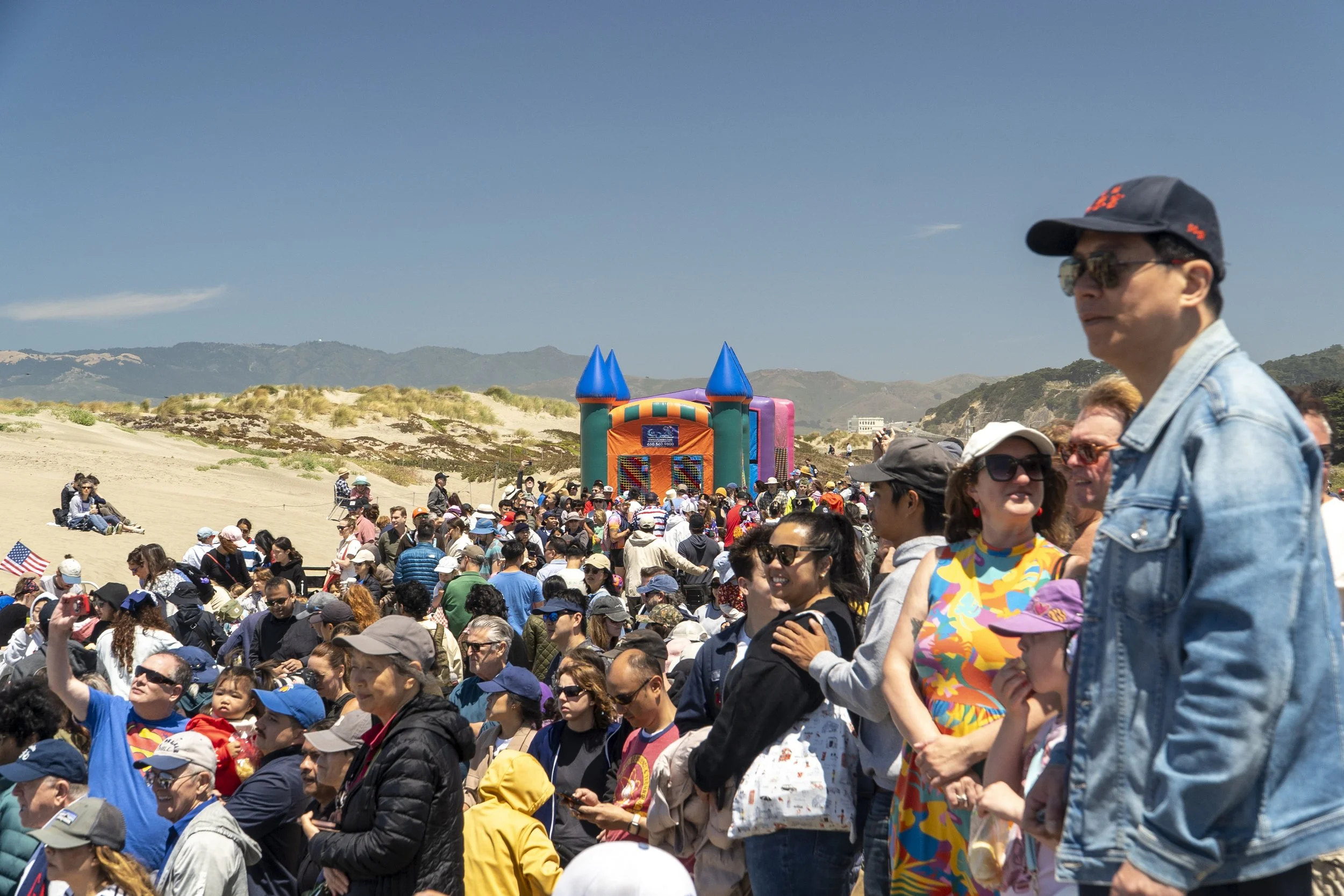 A large crowd of people gathered outdoors on a sunny day, with a colorful inflatable castle in the background and mountainous landscape beyond.