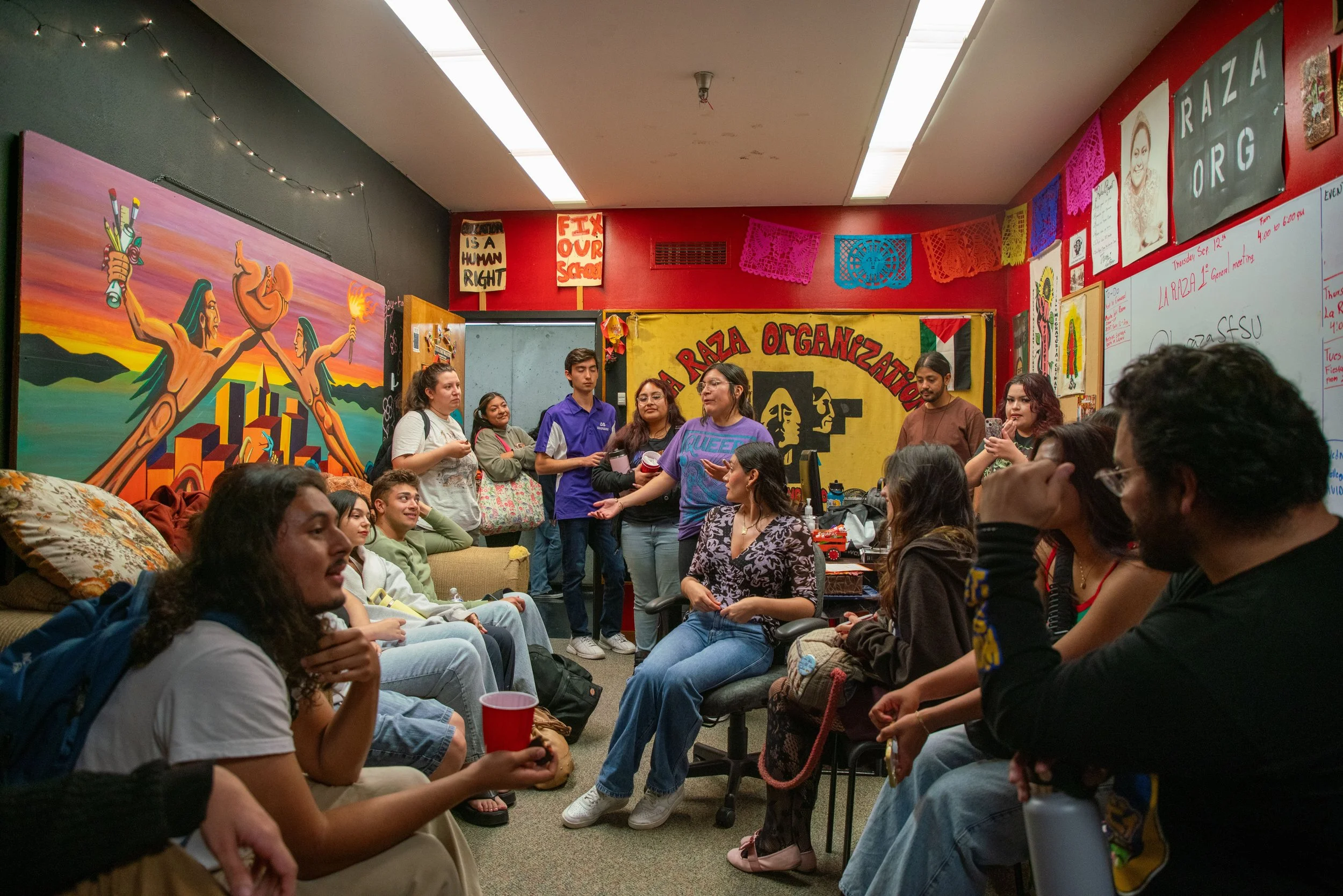 Group of young people gathered in a colorful room with murals and posters, engaged in conversation and activities, suggestive of a community or cultural event.