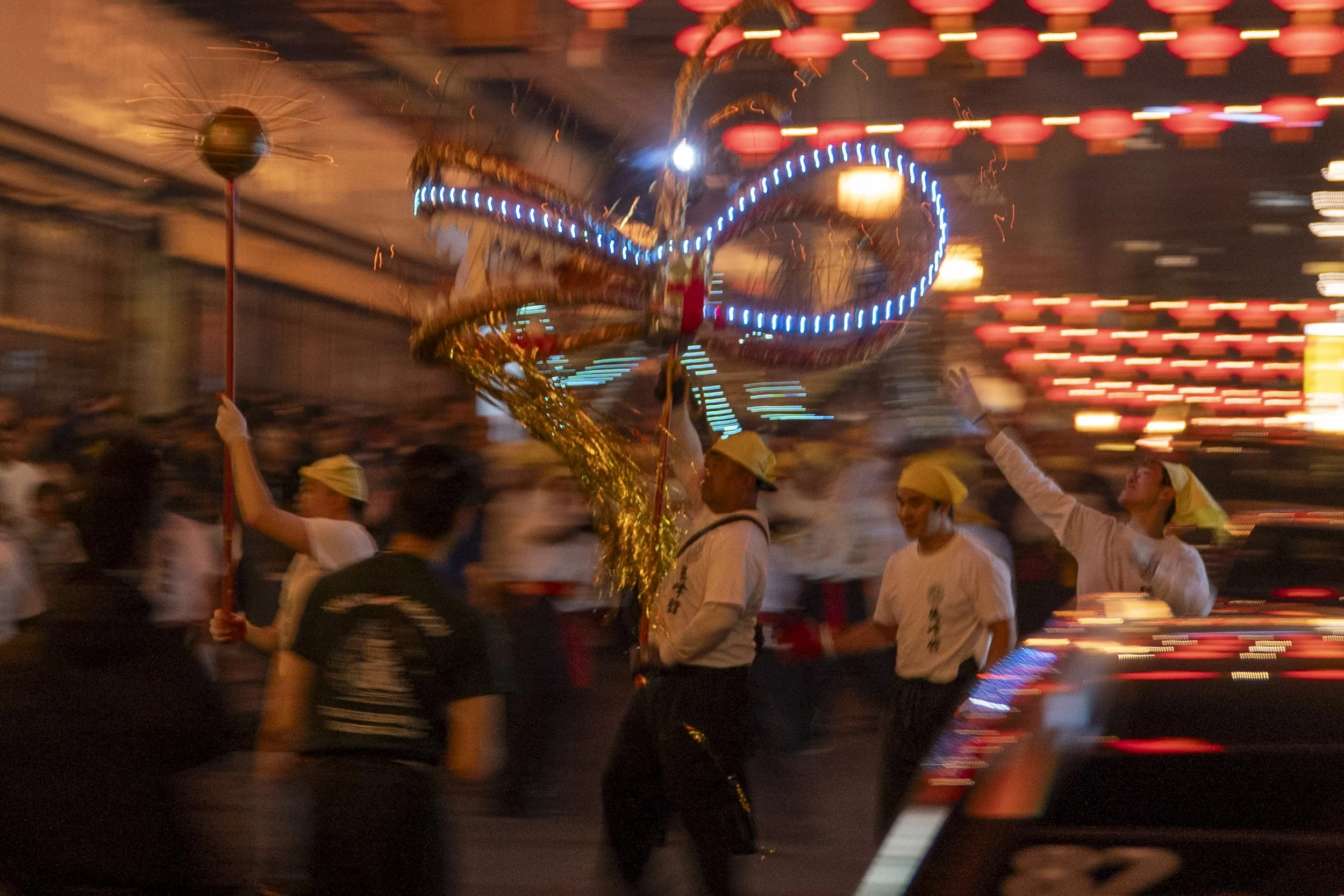 A nighttime scene of a parade featuring a person holding a flashing dragon-shaped lantern with blue lights, surrounded by dancers wearing white shirts and yellow headscarves, with a background of colorful illuminated lanterns.
