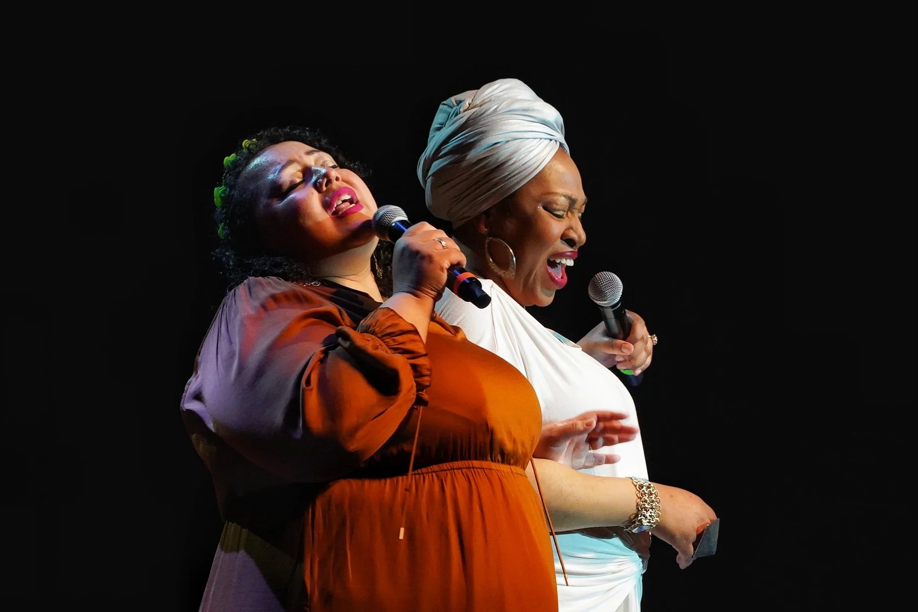Two women singing passionately on stage, holding microphones, against a black background.