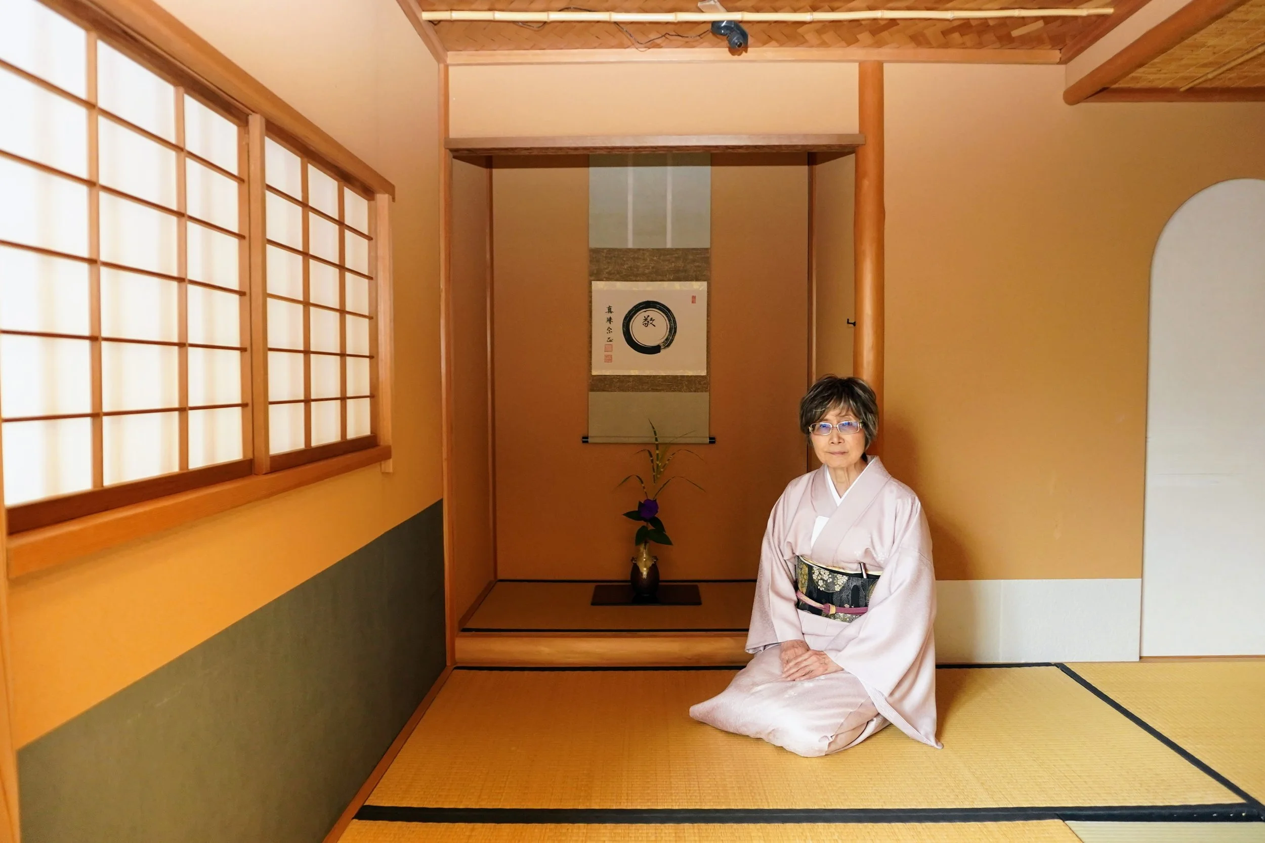 A woman in traditional Japanese clothing sits on tatami mats in a Japanese room with shoji screens and a tokonoma alcove.