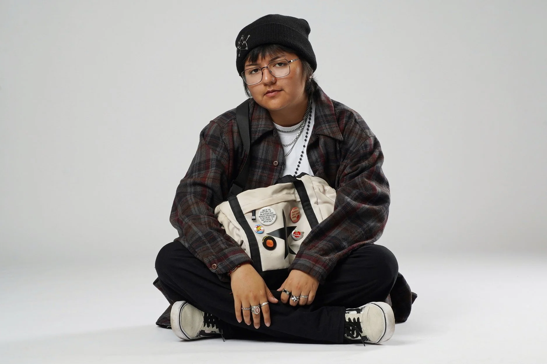 Person with short dark hair, glasses, wearing a black beanie, plaid shirt, black pants, and white sneakers, sitting cross-legged on the floor with a white backpack decorated with pins.