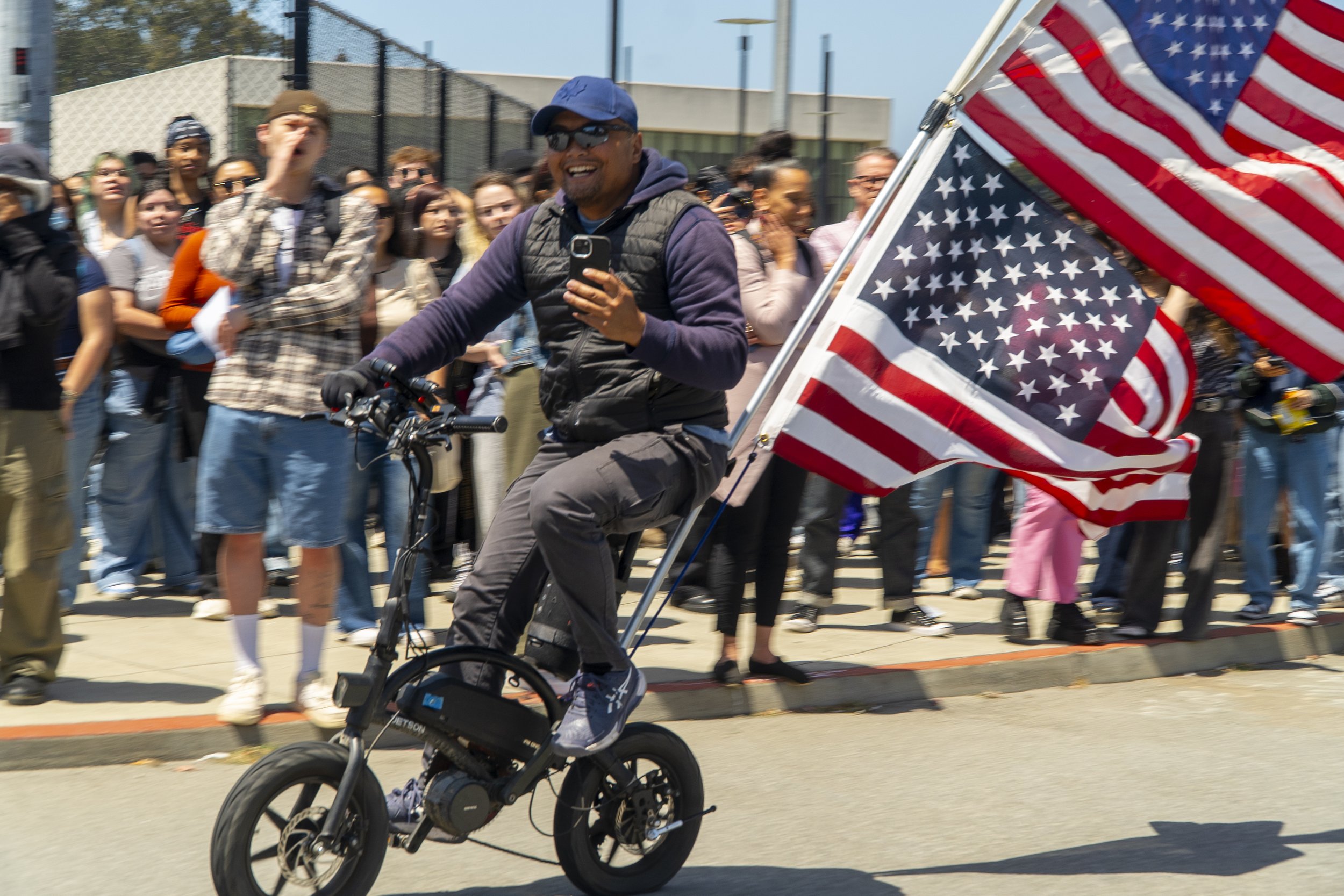 A man riding a small electric bike with American flags attached, smiling and looking at his phone, with a crowd of people watching in the background on a sunny day.