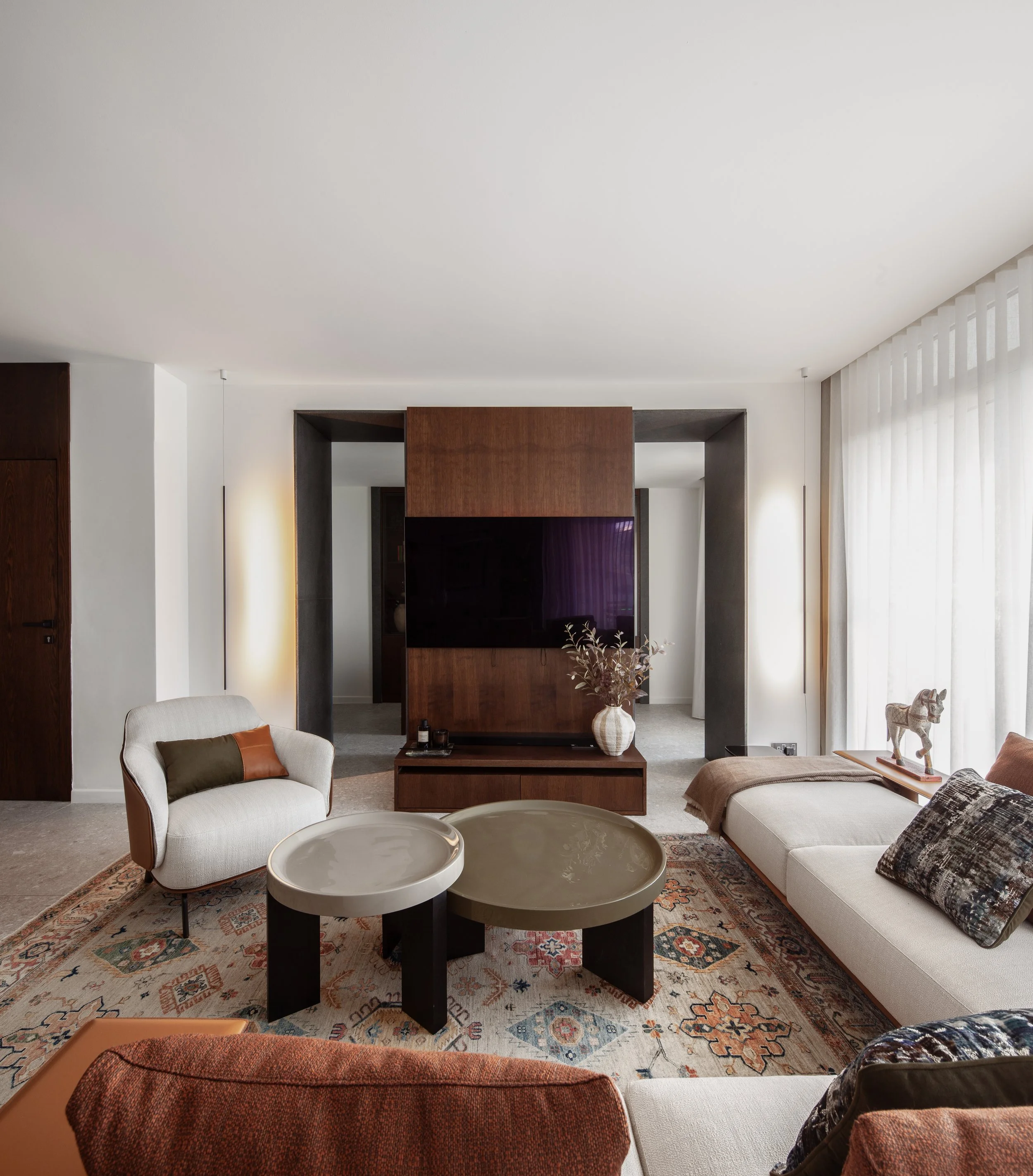 Modern living room with beige sofas, circular coffee tables, a patterned rug, a large flat-screen TV, and floor-to-ceiling curtains