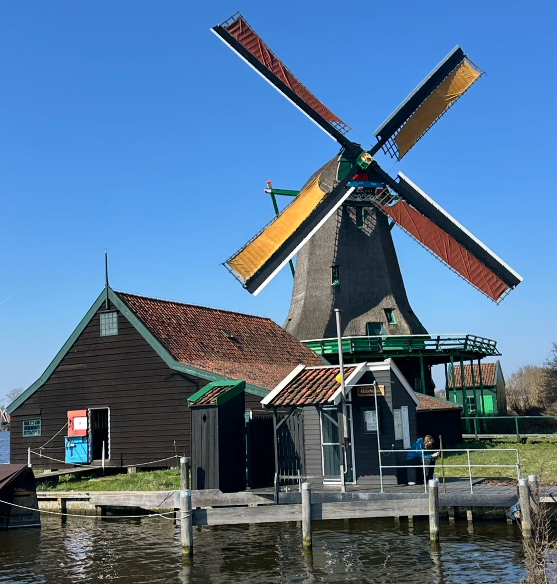 A traditional Dutch windmill with four sails on a waterfront, accompanied by nearby small buildings and a parasol, under a clear blue sky.