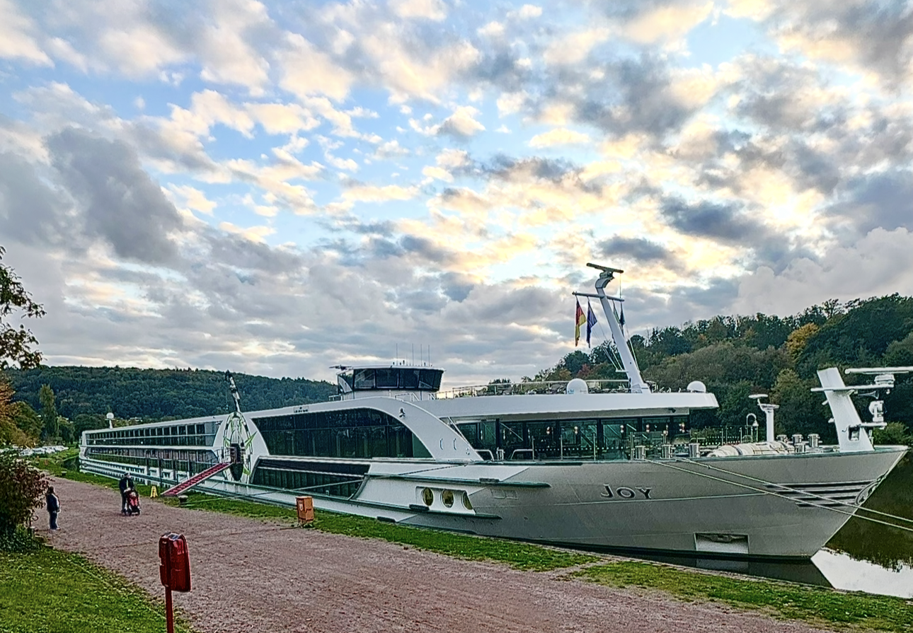 Large luxury white boat named JOY docked beside a grassy path on a cloudy day with hills in the background.