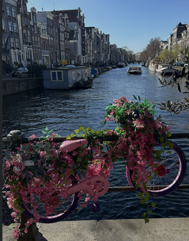 A canal in an urban area with tall buildings on both sides, boats docked along the water, and a pink bicycle decorated with pink flowers hanging on a railing in the foreground.