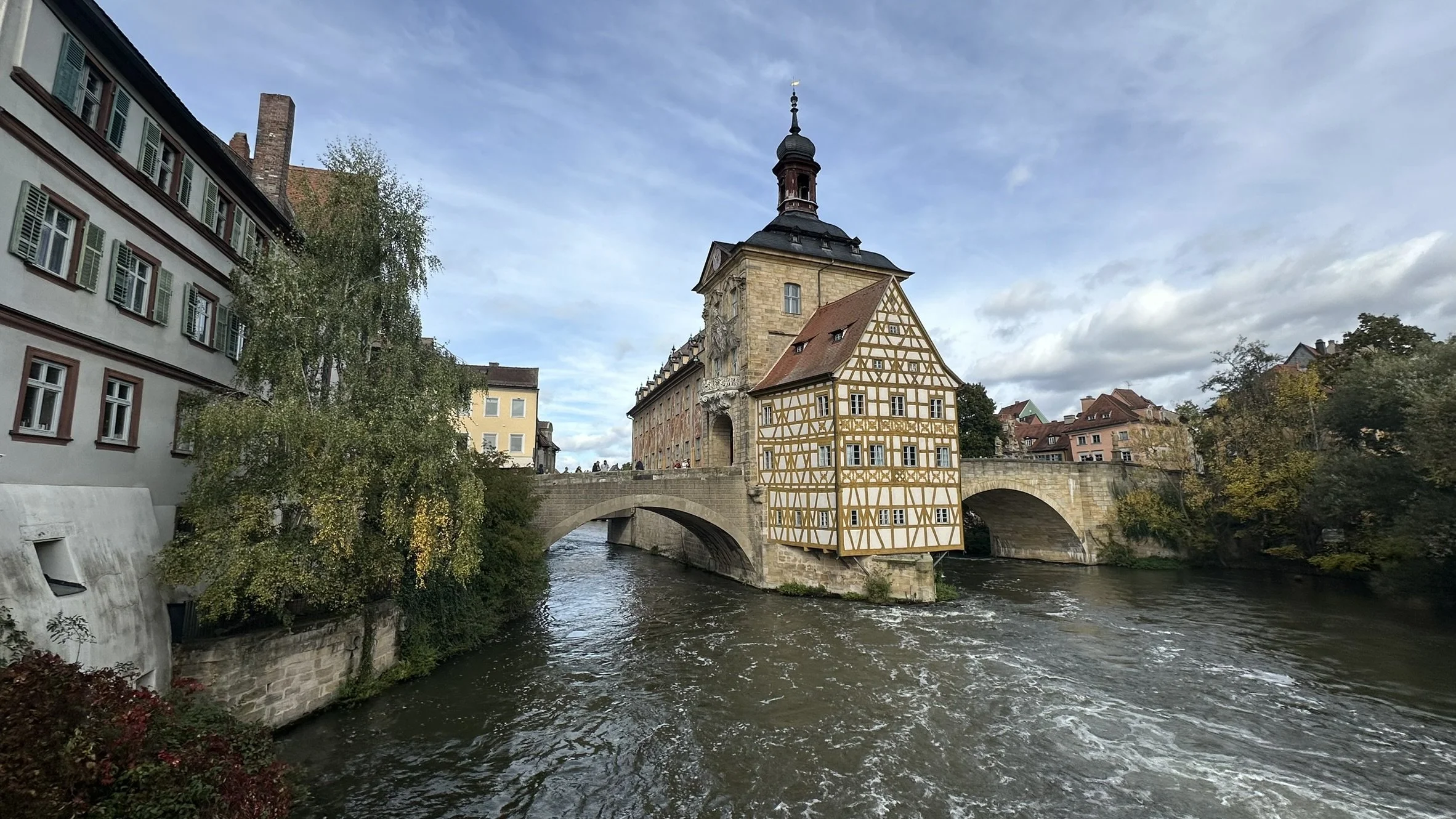 The image features a historic bridge over a river with a building that has a half-timbered facade and a tower with a spire behind it. There are trees on either side of the river and residential buildings in the background under a partly cloudy sky.