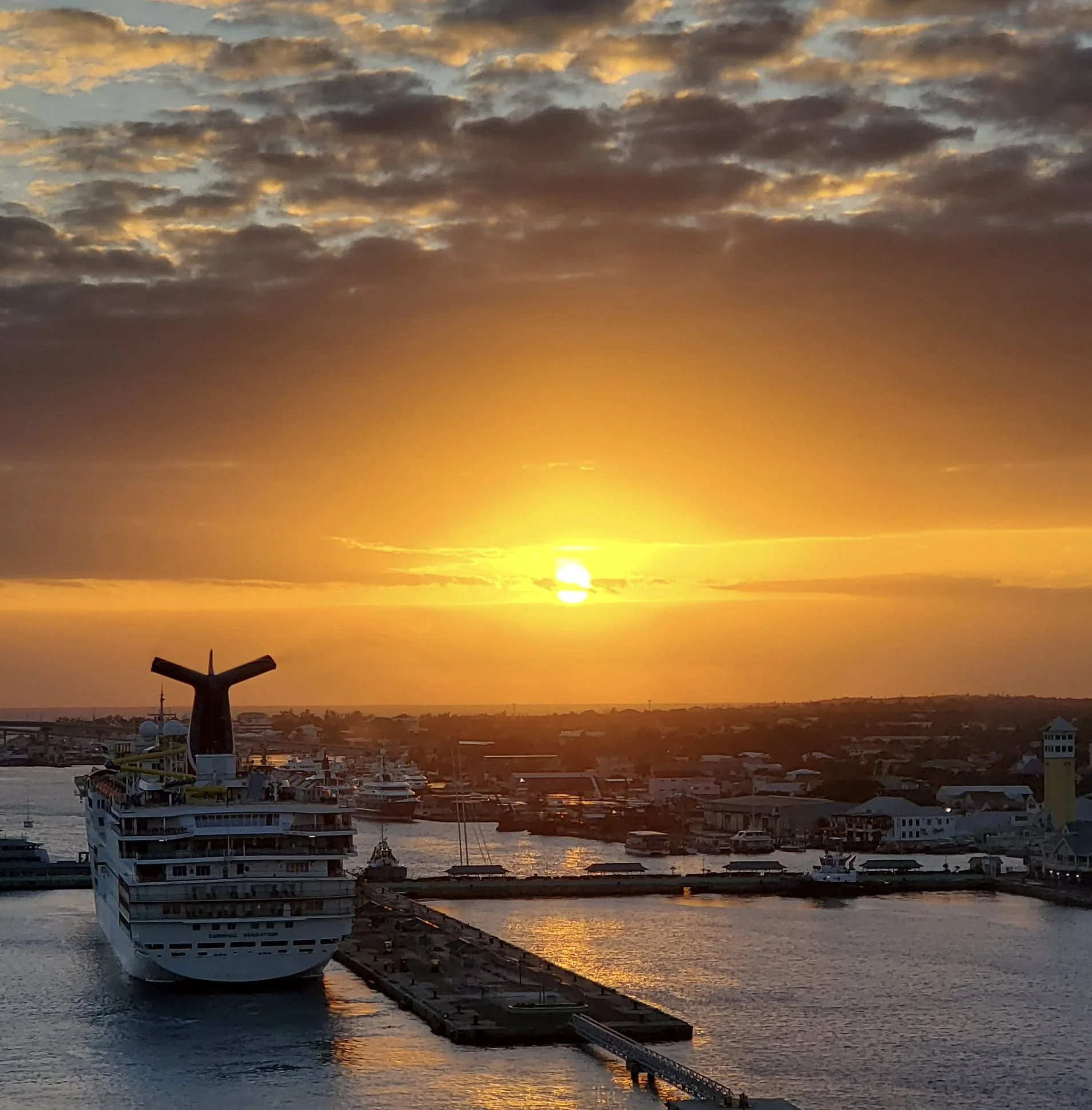 A large cruise ship docked at a harbor during sunset, with clouds in the sky and water reflecting the golden hues of the setting sun.