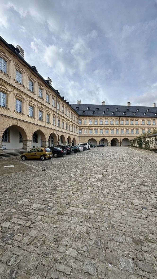 A cobblestone courtyard surrounded by a historic European-style building with arched doorways and multiple windows, with parked cars along the side and a cloudy sky above.