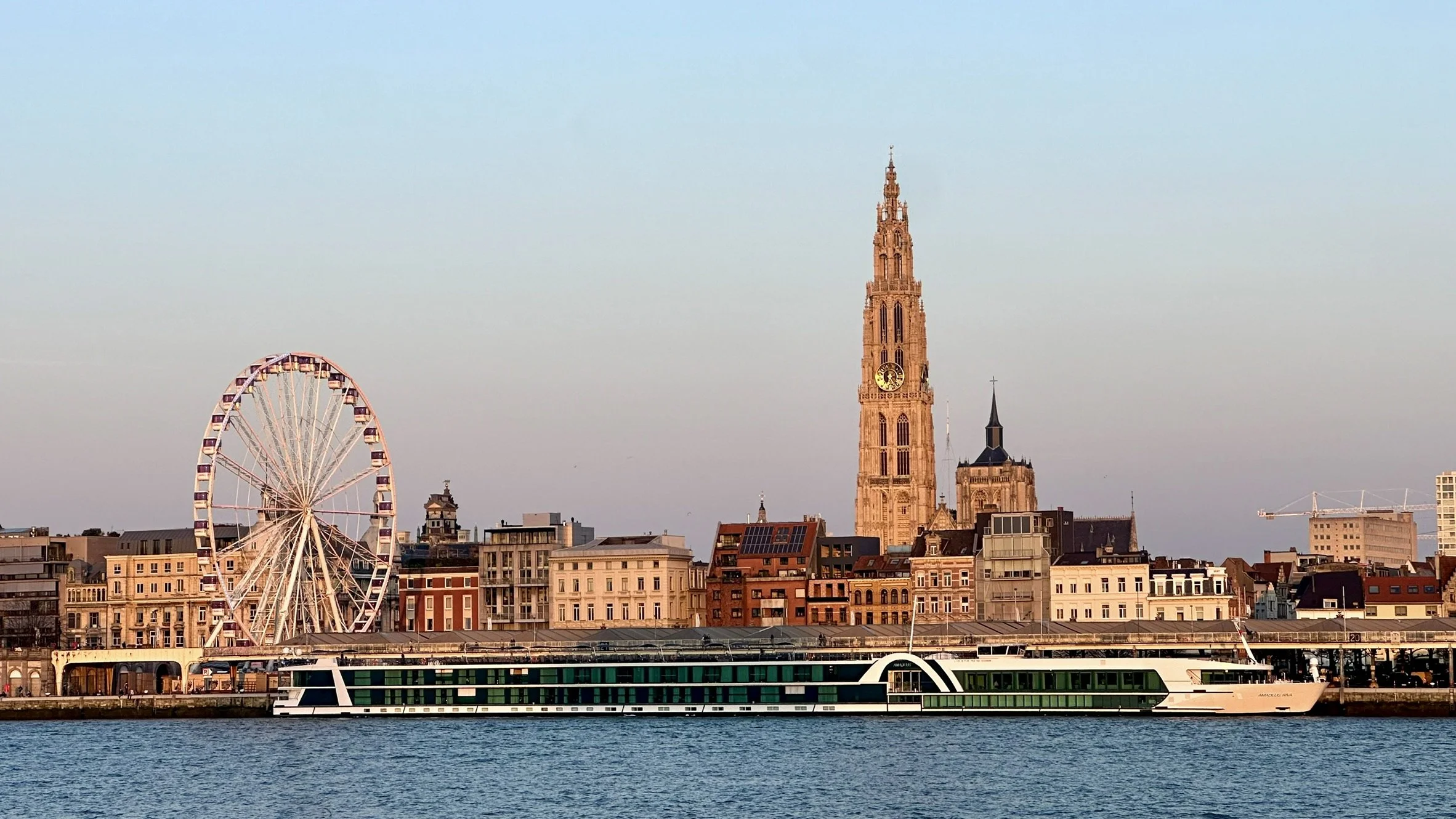 City skyline with a large Ferris wheel, historic church with a tall clock tower, modern buildings, and a boat on the water in the foreground.