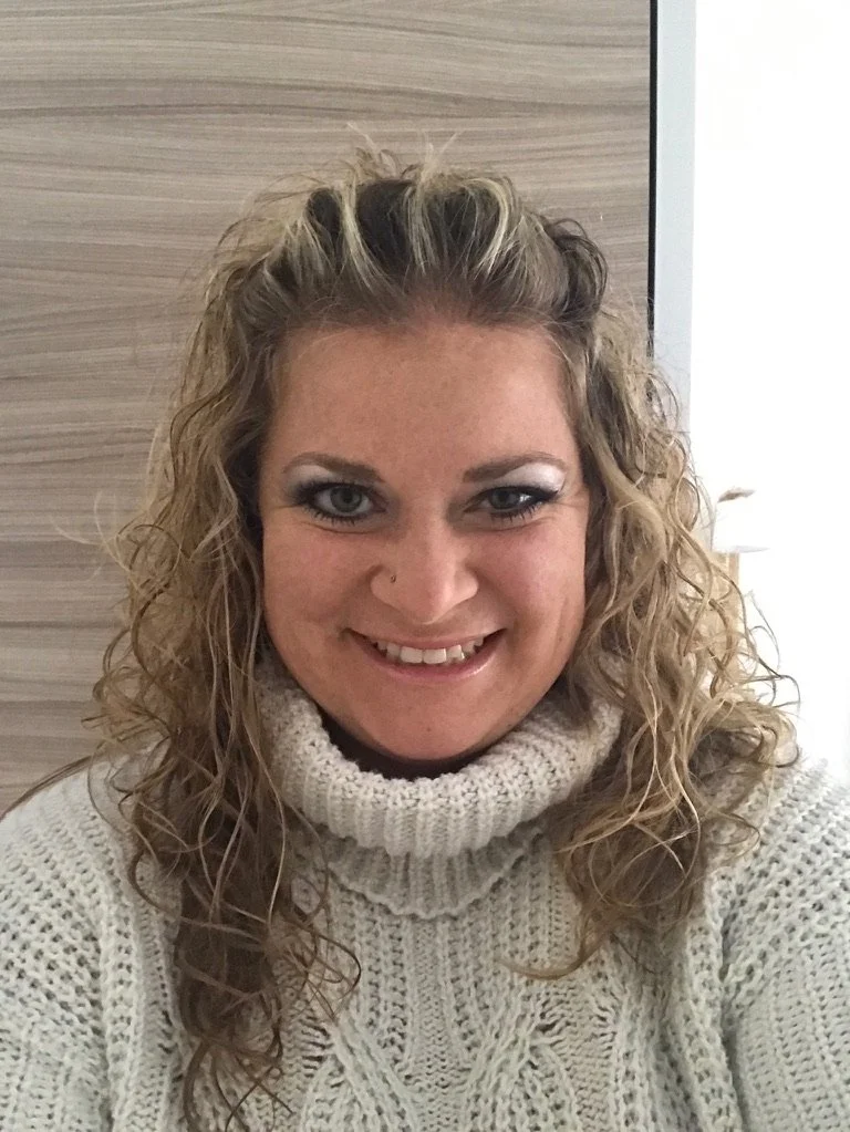 A woman with curly blonde hair and blue eyes wearing a white knit turtleneck sweater, smiling at the camera indoors.