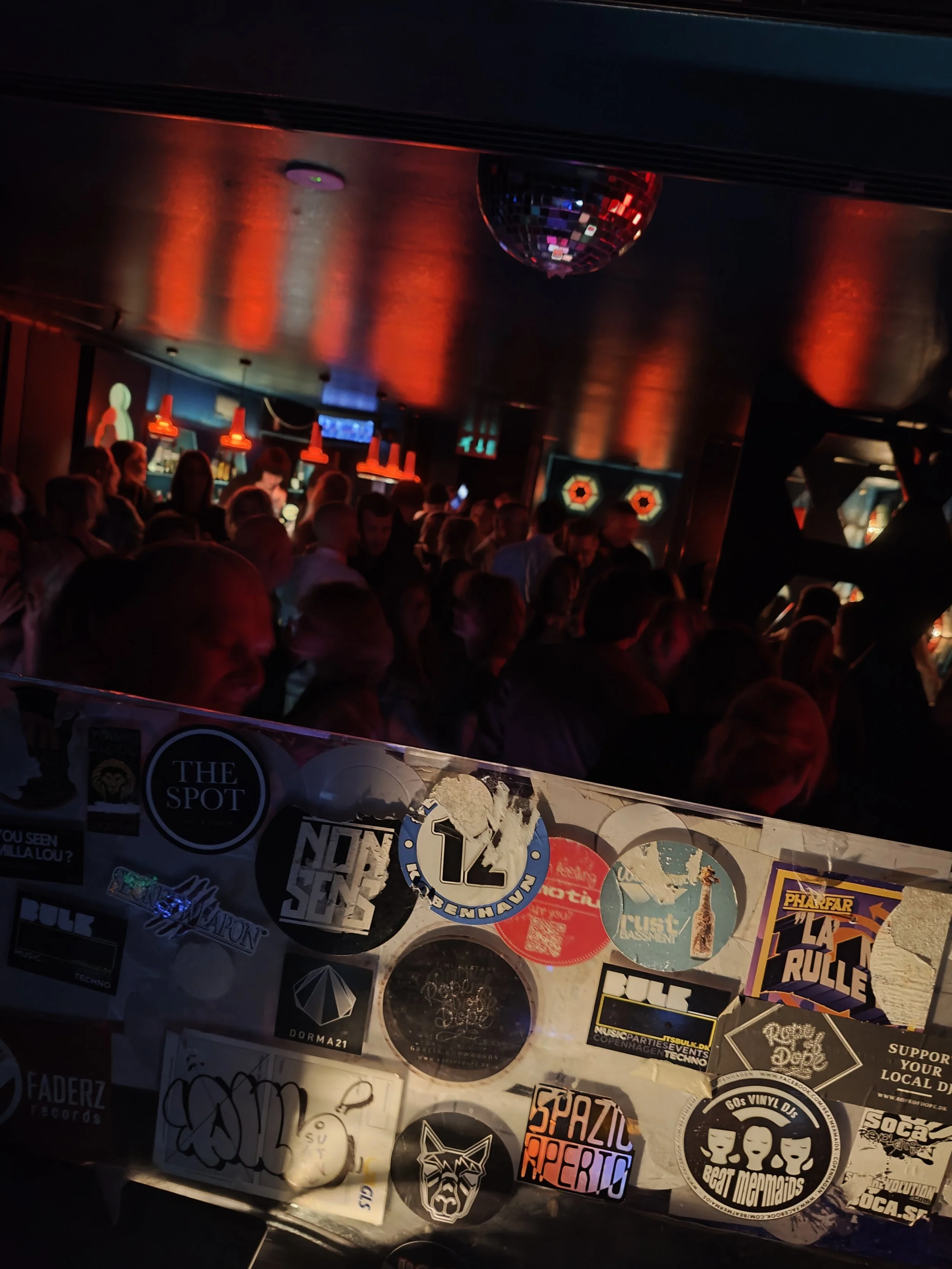 En fyldt natklub med rødt og blåt lys, en diskokugle hængende fra loftet og forskellige klistermærker på en overflade i forgrunden.