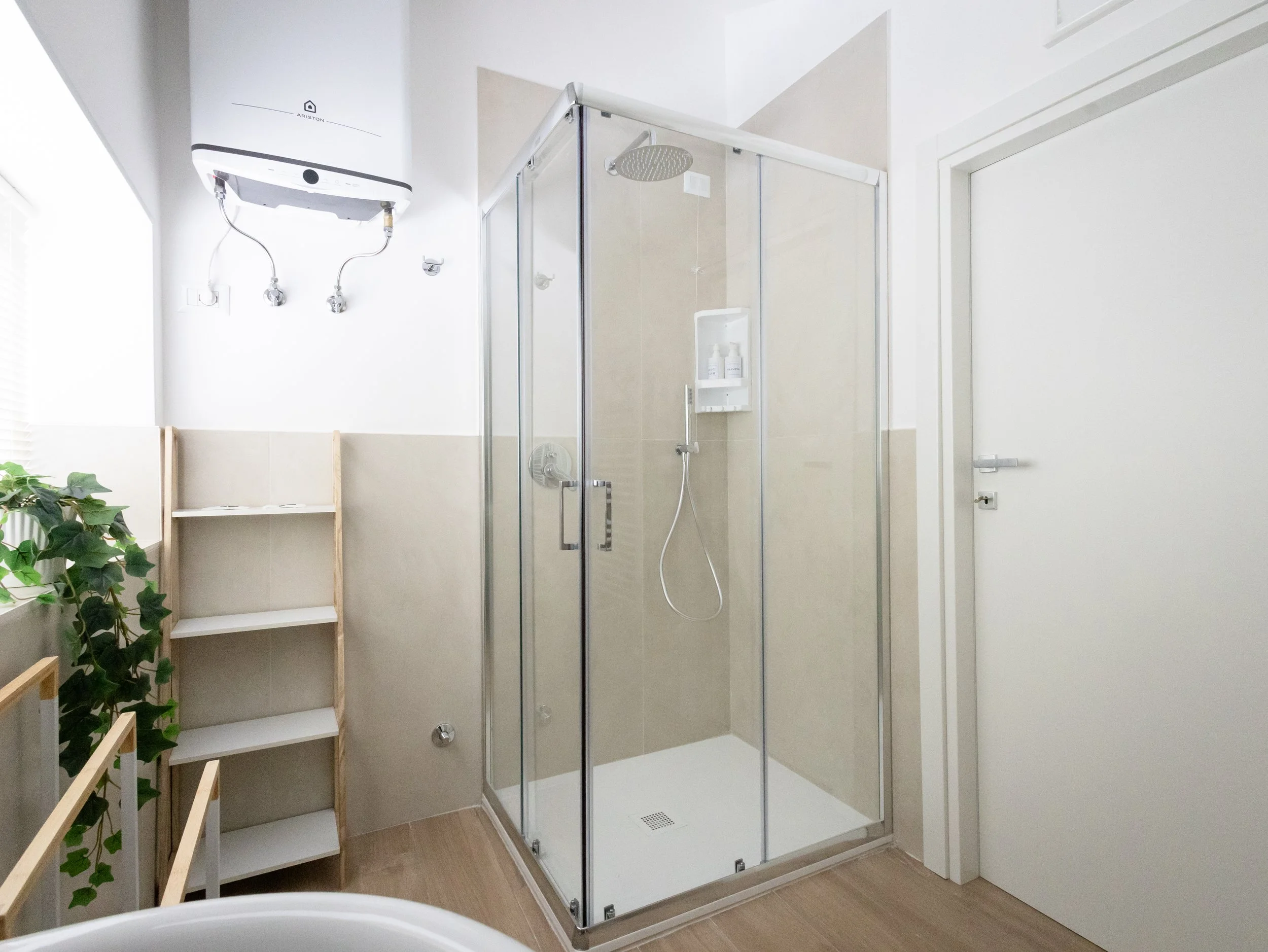 Doccia con pareti in vetro, rubinetto e soffione, situata in un bagno moderno con pareti bianche, una pianta verde sul lato sinistro e una scaffalatura di legno vuota.