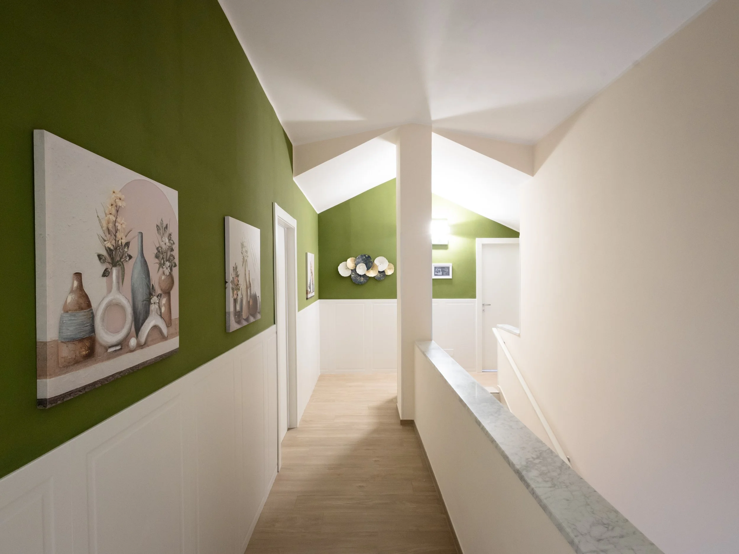 Interno di una casa con pareti verde oliva e bianche, quadri appesi e una pianta decorativa sopra un mobile.