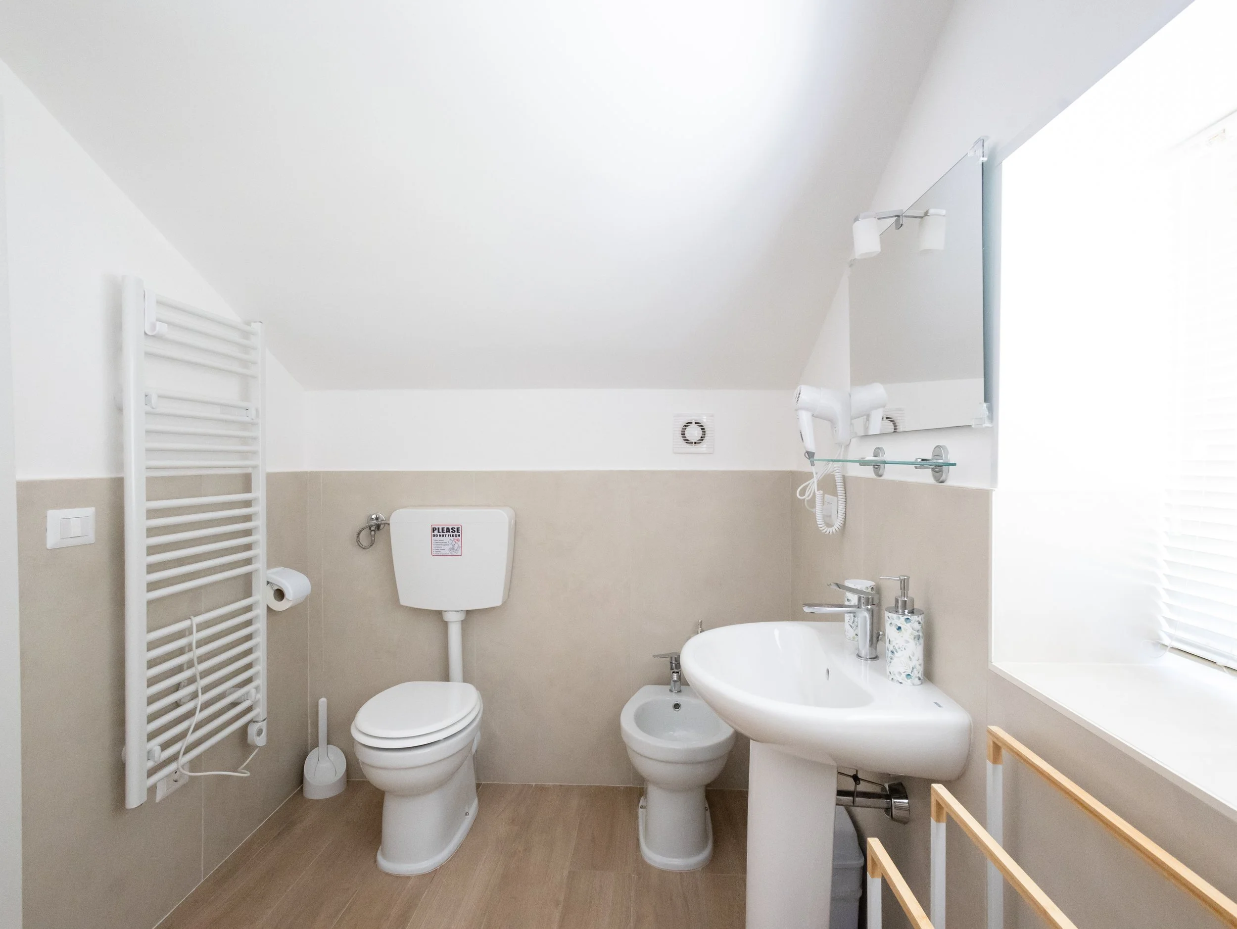 Bagno moderno con doppi sanitari, lavabo e specchio, scaldasalviette bianco e finestra luminosa.