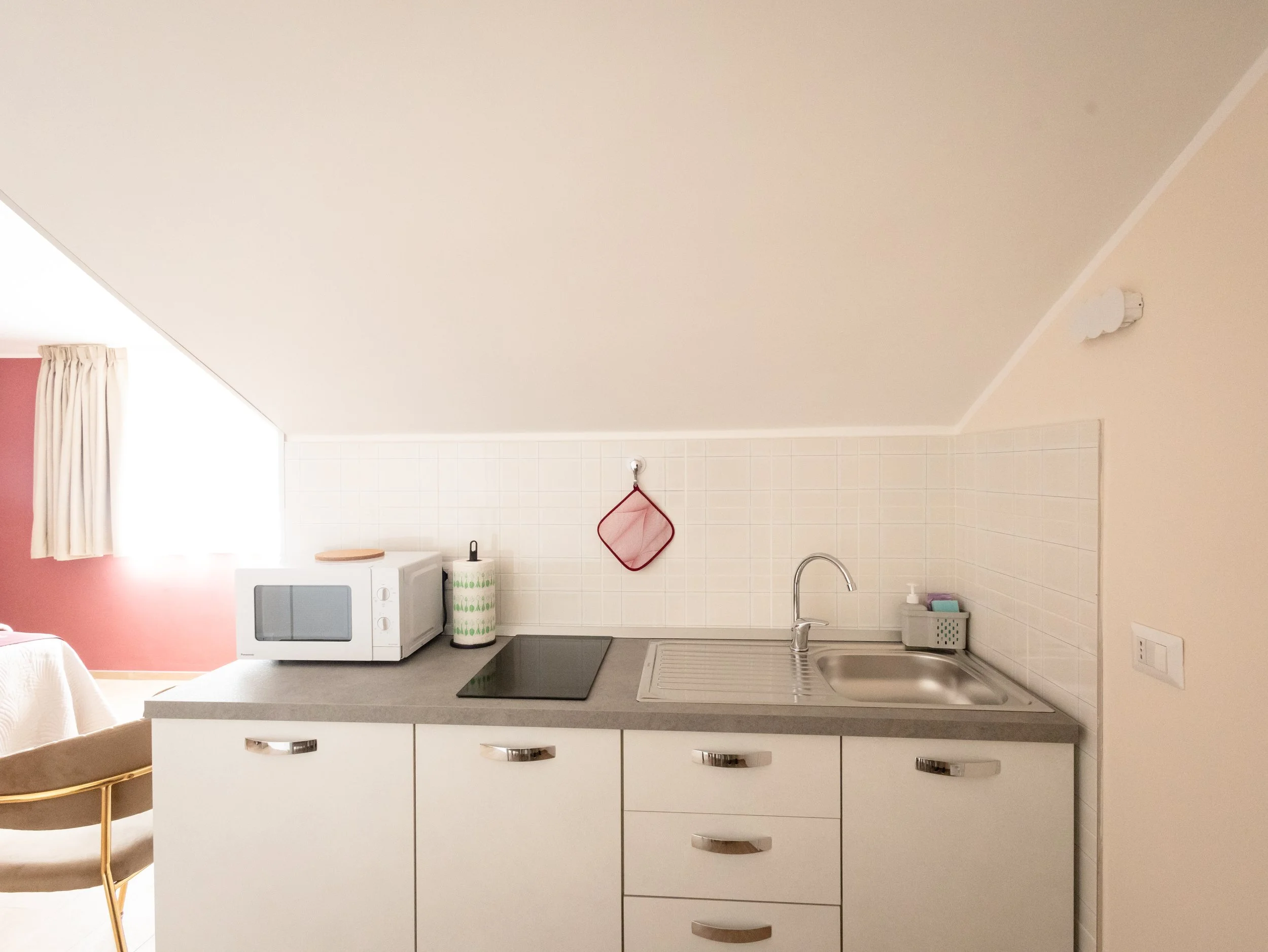 Cucina con lavabo, forno a microonde, piani di lavoro, pareti rivestite di piastrelle chiare, e una finestra con tendina bianca. C'è anche un mobile con cassetti bianchi e maniglie argentate, un asciugamano a fiore, e un utensile in una ciotola accan