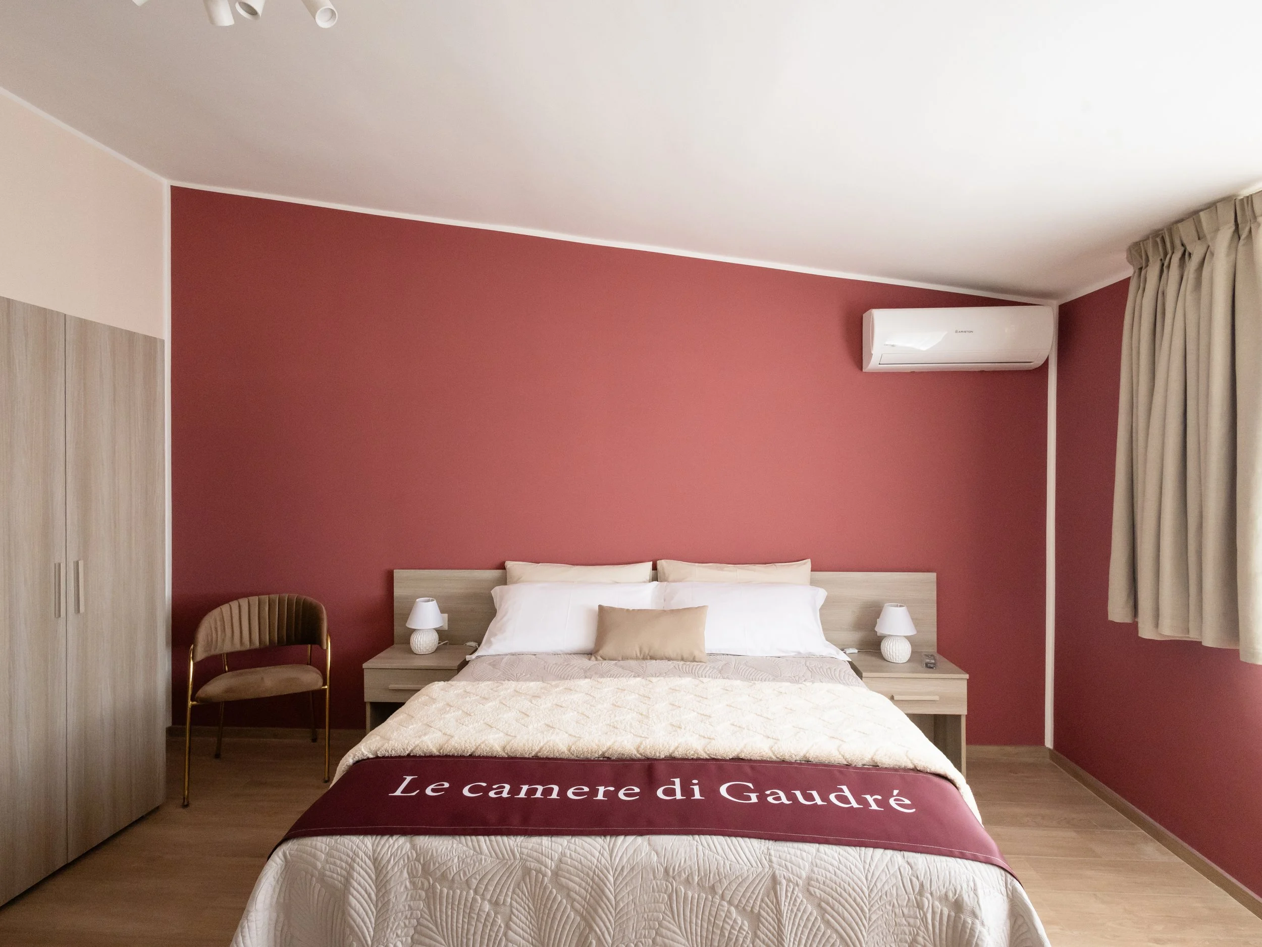 Camera da letto con parete rossa, letto con testata in legno, cuscino beige, biancheria bianca, due lampade ai lati del letto, sedia in velluto beige e armadio in legno chiaro. Tende beige e aria condizionata bianca