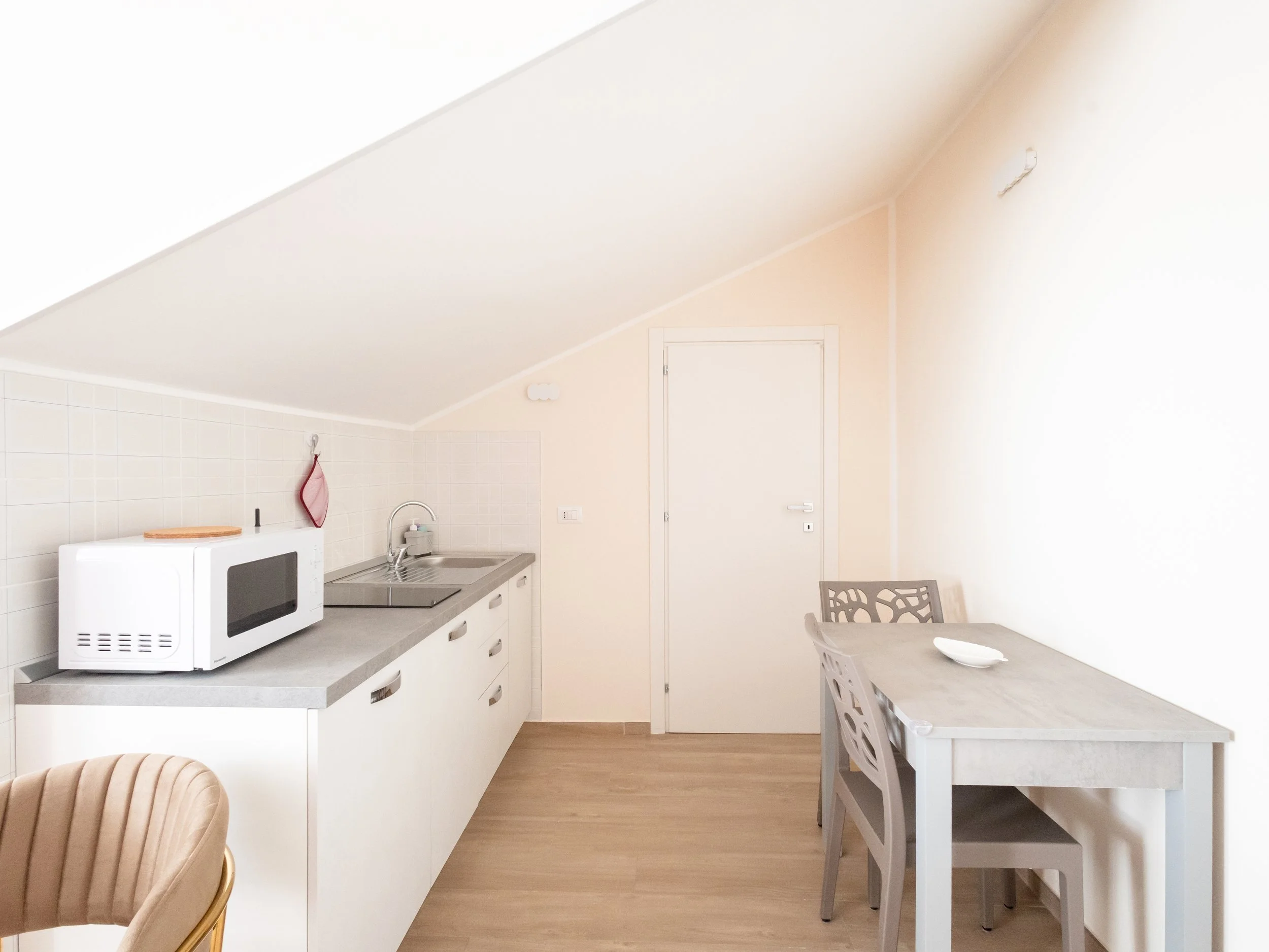 Cucina con mobile bianco, microonde, lavandino, tavolo con sedie grigie, pareti beige, porta bianca, parete con piastrelle bianca, soffitto inclinato, pareti lisce