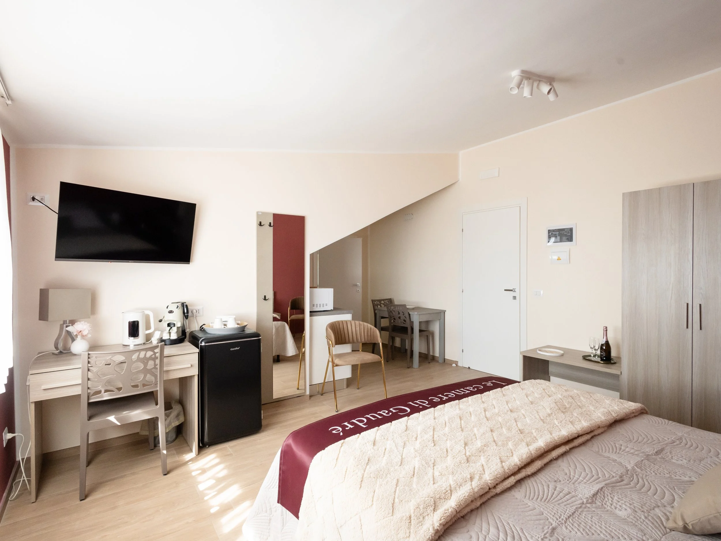 Interno di una camera da letto con pareti beige, un televisore a parete, una scrivania con una sedia, un letto con coperta beige e un pigiama, e una parete con specchio