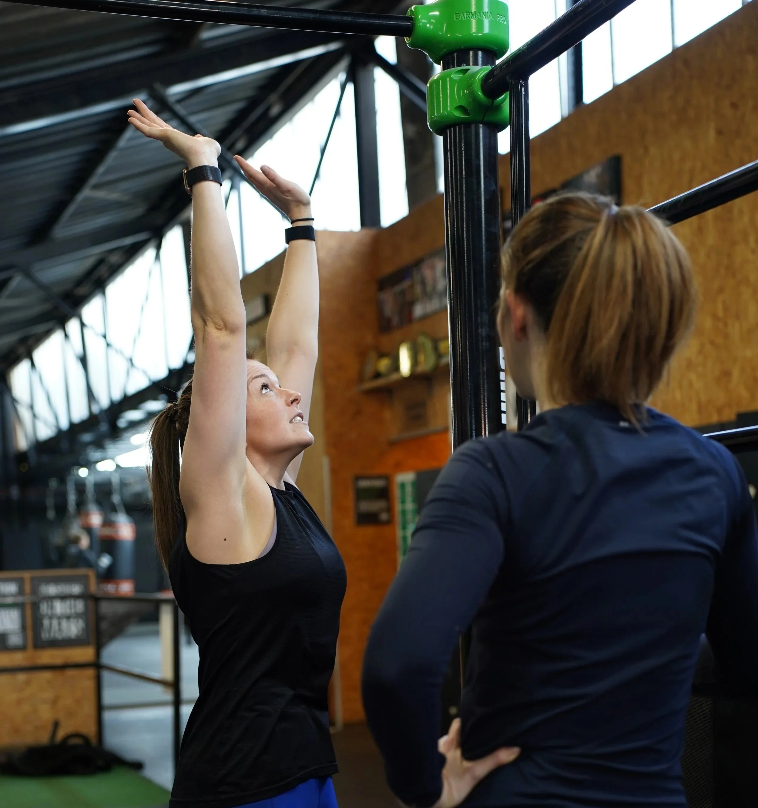 A personal trainer in a gym instructing her female client on how to perform an exercise using a pull-up bar.