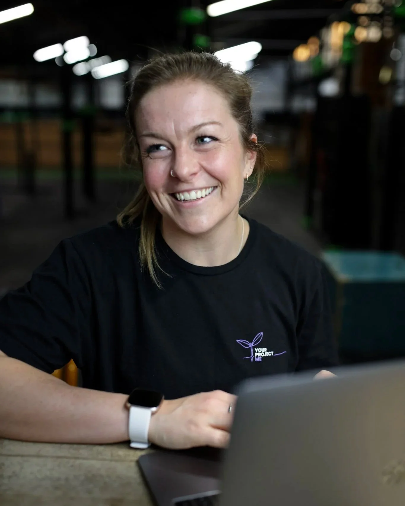 Smiling female coach with a nose piercing, wearing a black T-shirt with a small business logo that says 'Your Project Me,' sitting at a table with a laptop, to represent online coaching.