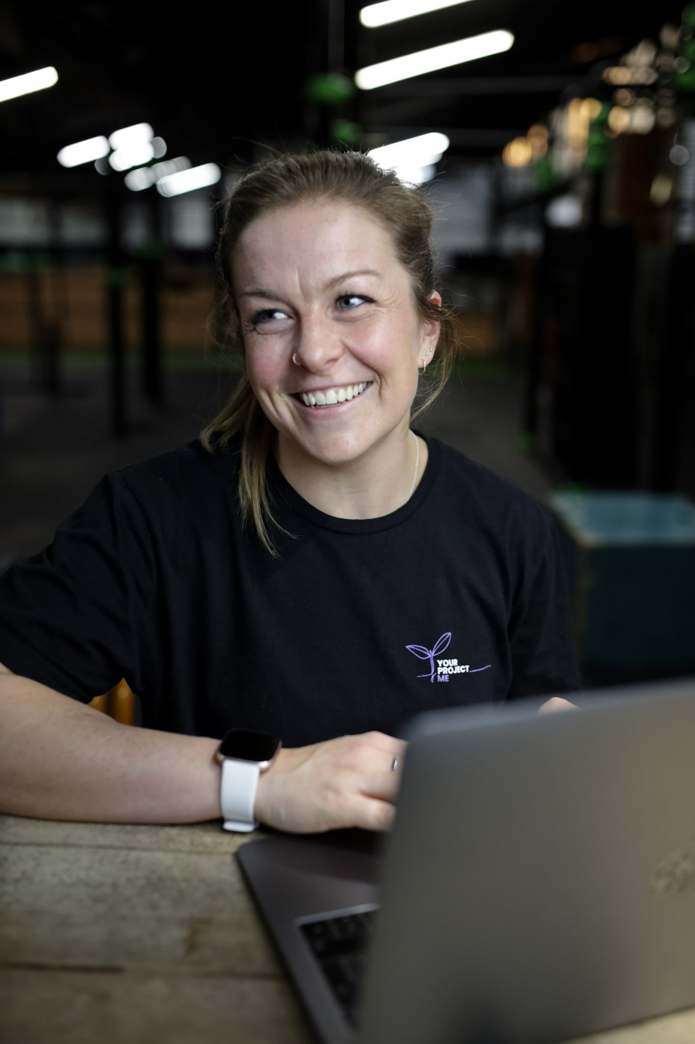 A smiling female coach wearing a black T-shirt with a business logo, sitting at a table with a laptop, to represent online coaching.