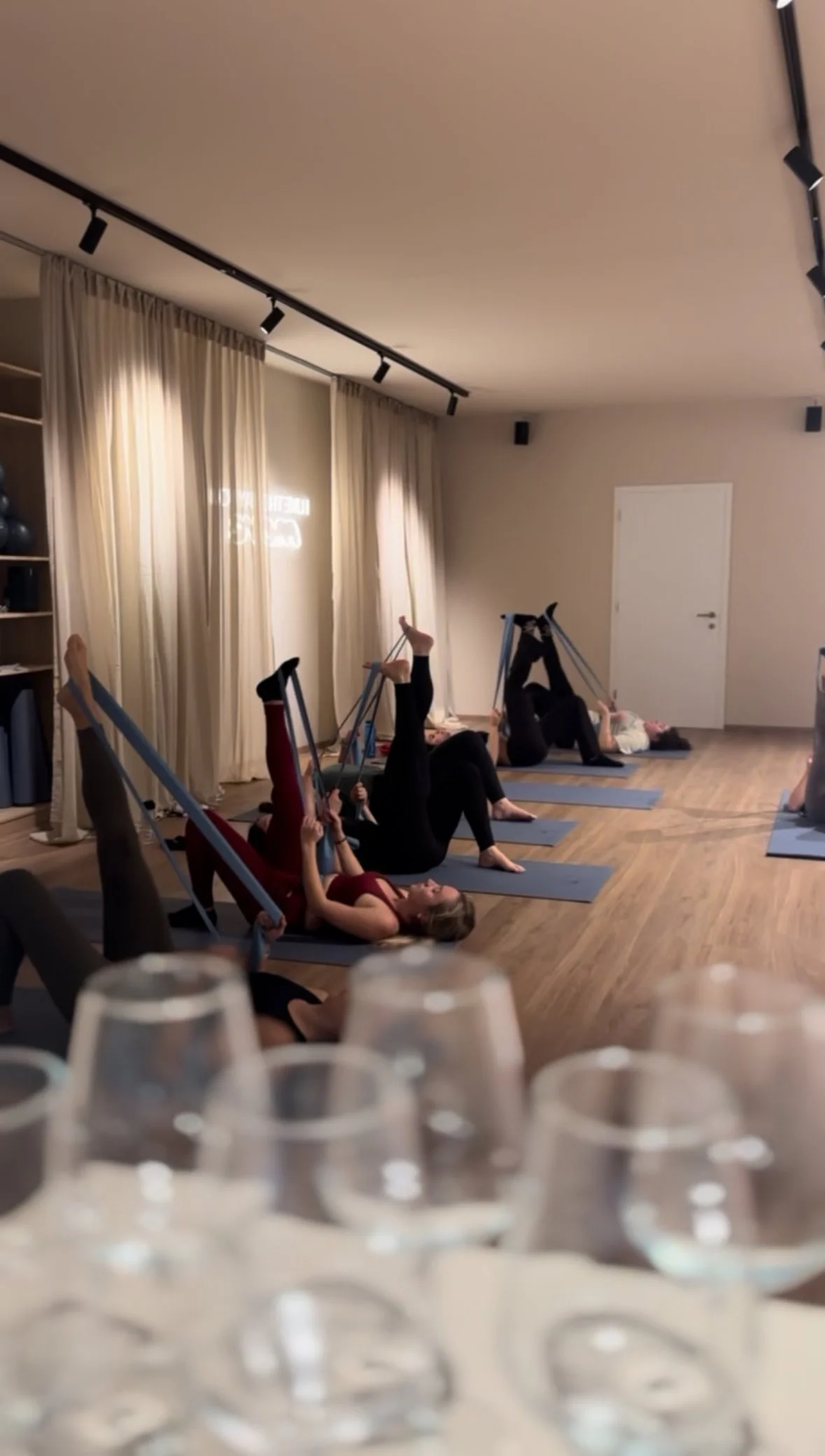 Menschen, die in einem Fitnessstudio Yoga mit Schlingentrainern machen, während im Vordergrund mehrere leere Weingläser stehen.