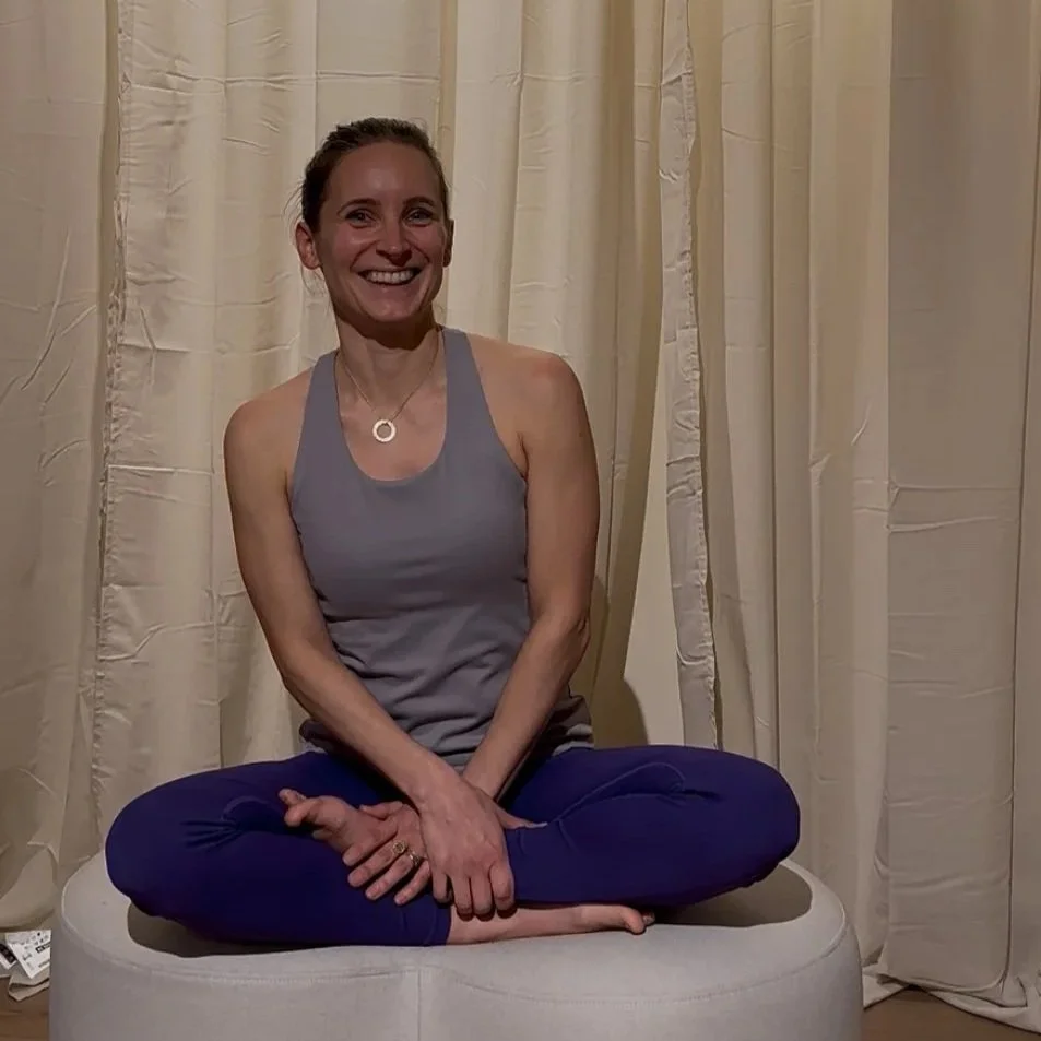 Eine lächelnde Frau sitzt im Schneidersitz auf einem runden Polster vor cremefarbenen Vorhängen.