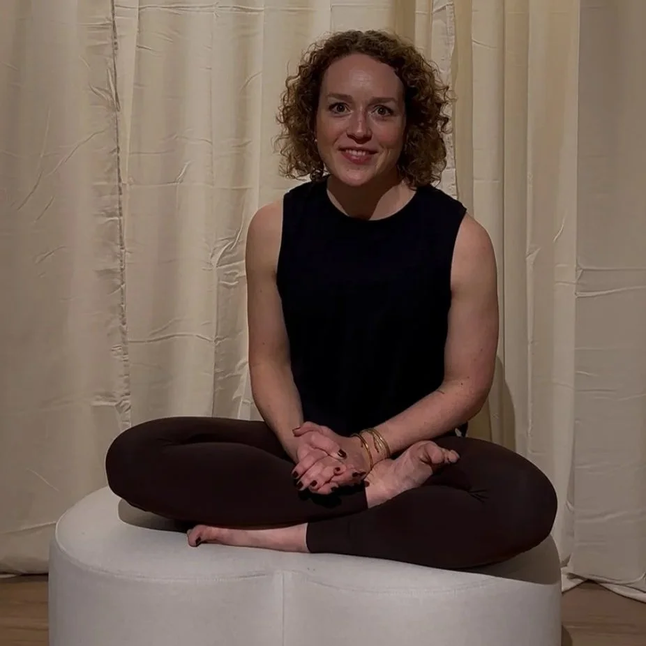 Eine Frau sitzt im Schneidersitz vor einem neutralen, hellen Vorhang und lächelt.