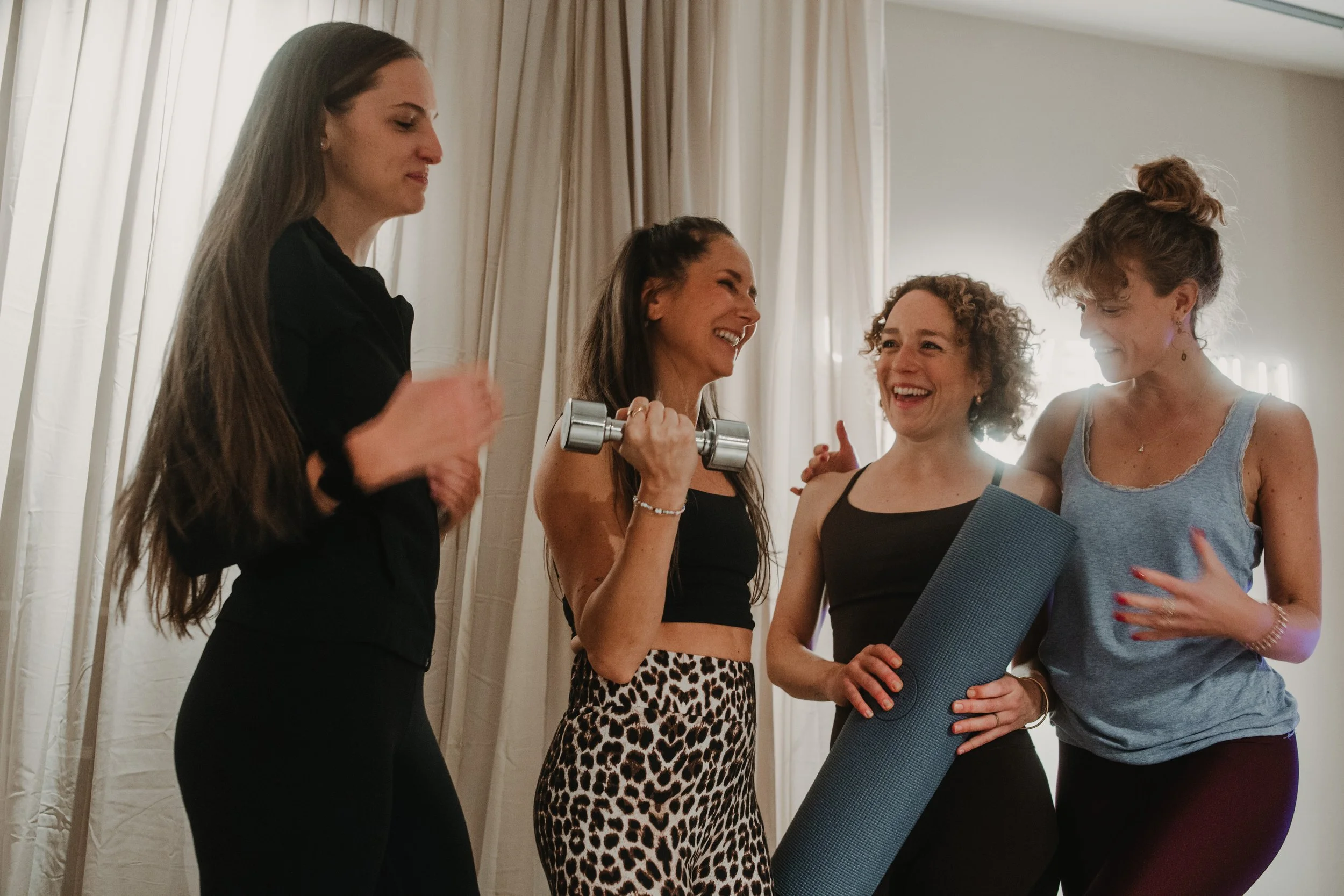 Vier Frauen beim Fitness, eine hält eine Hantel, eine andere eine Yogamatte, sie lachen zusammen.